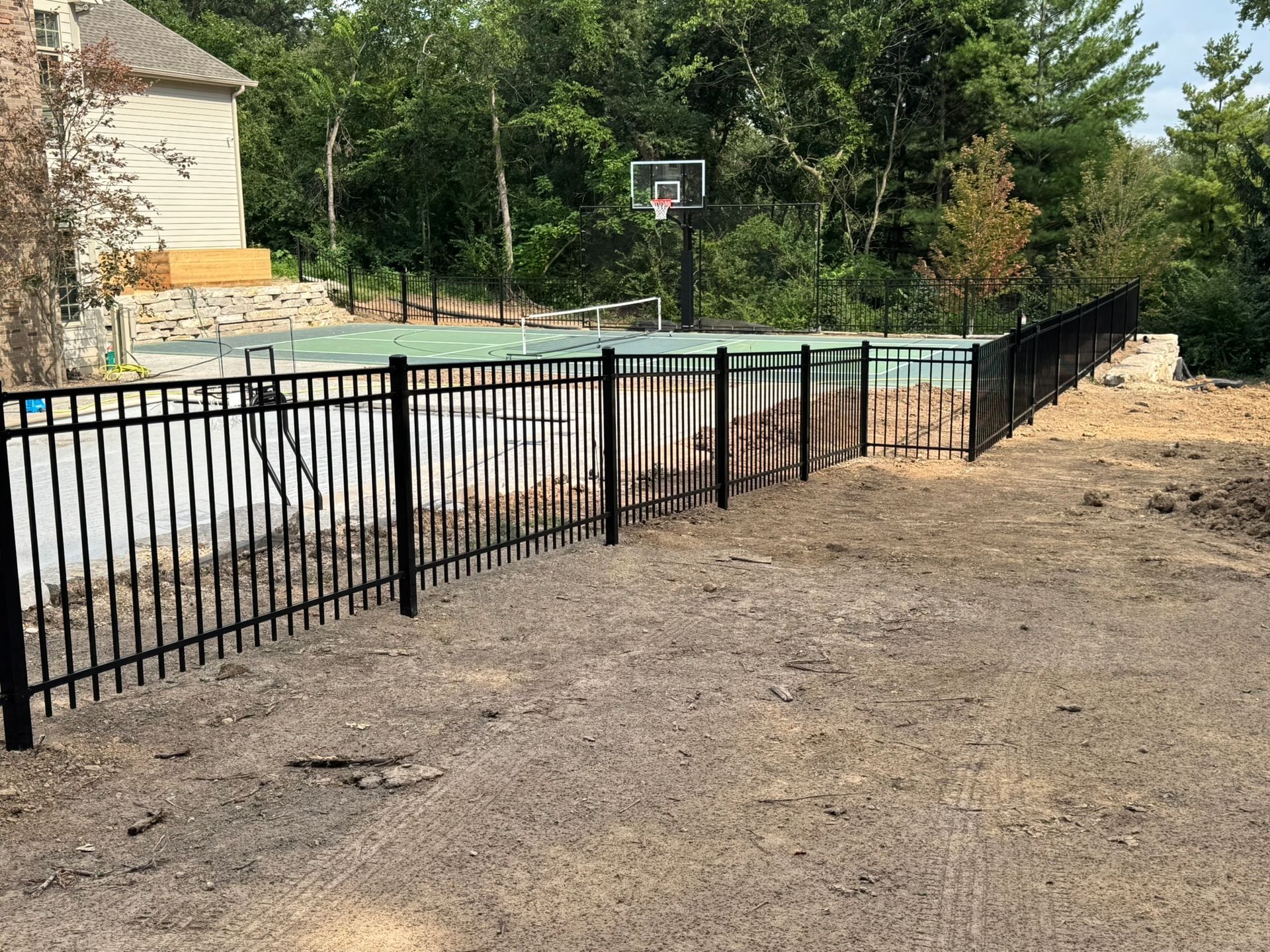 Black metal fence surrounds a backyard pool, adjacent to a house, basketball hoop visible in the distance.