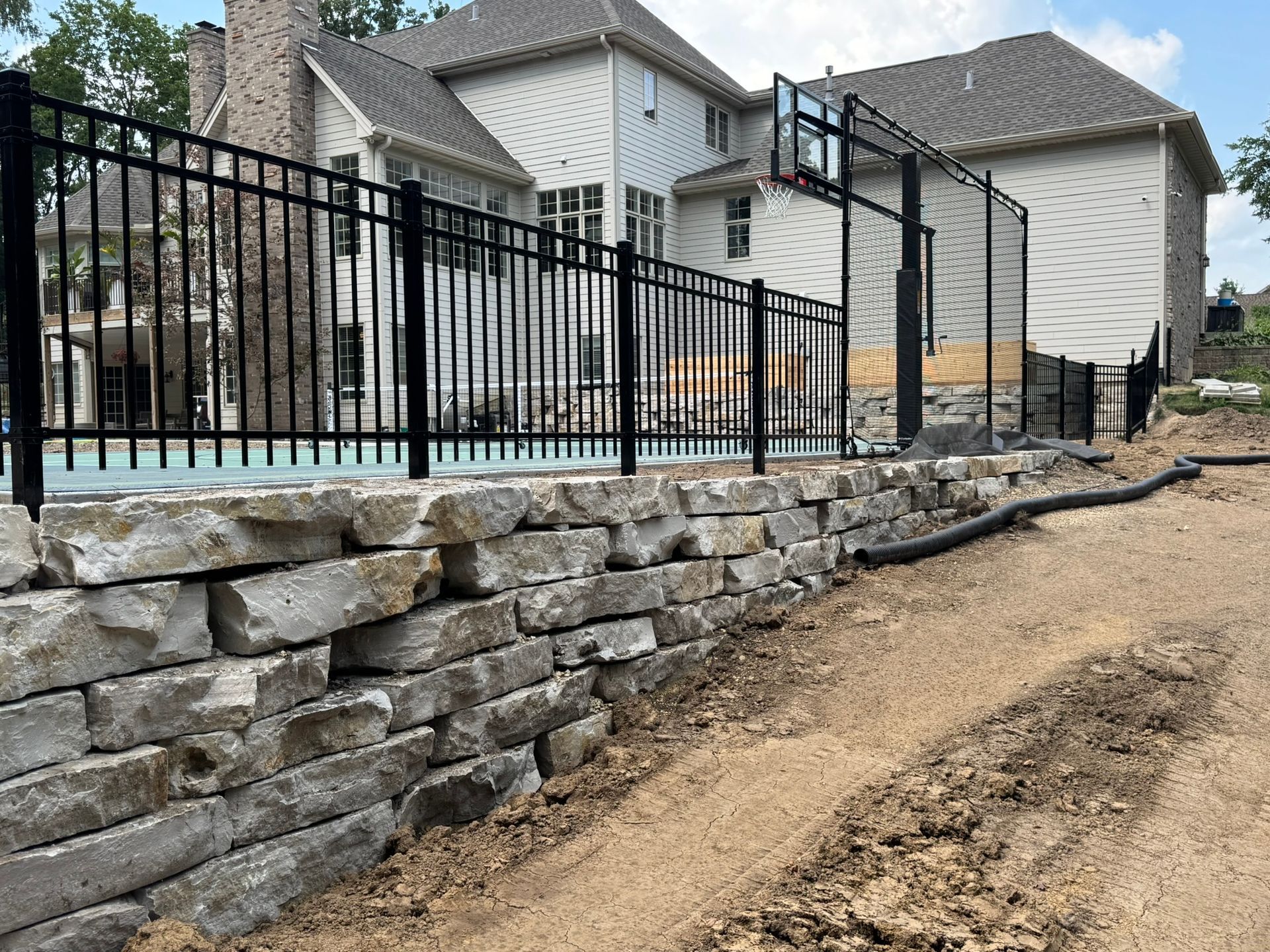 Black fence atop a stone retaining wall borders a pool and backyard of a large house.