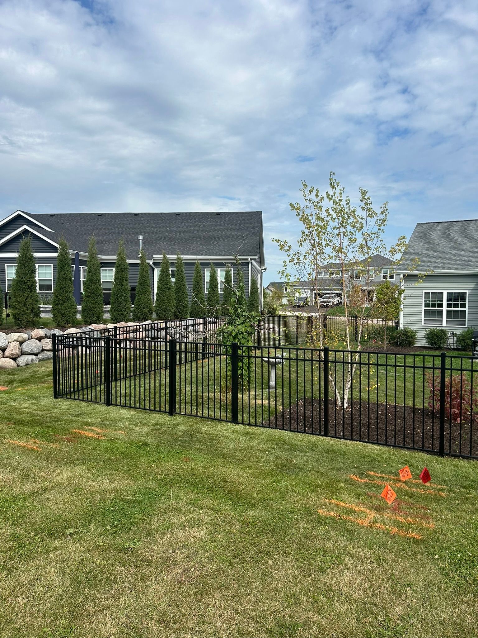 Black fenced backyard with green grass, small tree, and houses under a cloudy sky.
