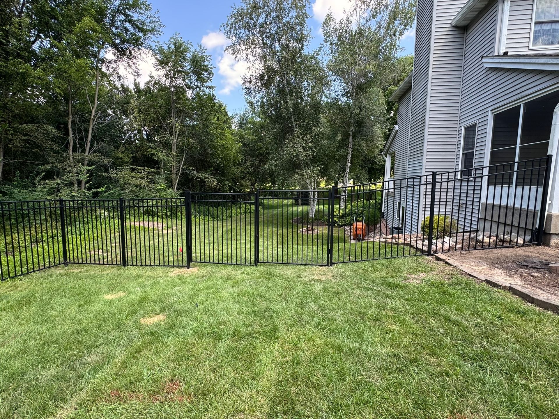 Black metal fence surrounding a grassy backyard next to a house with trees and a bright sky.