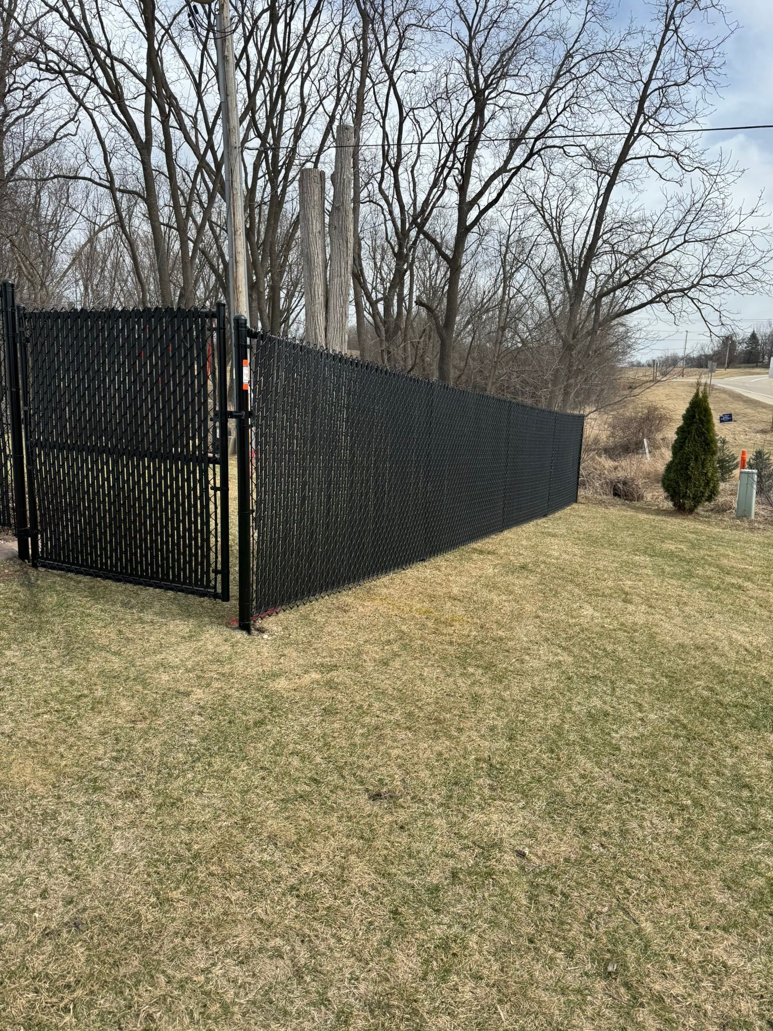 Black fence with privacy screening on grassy area. Trees and sky in the background.