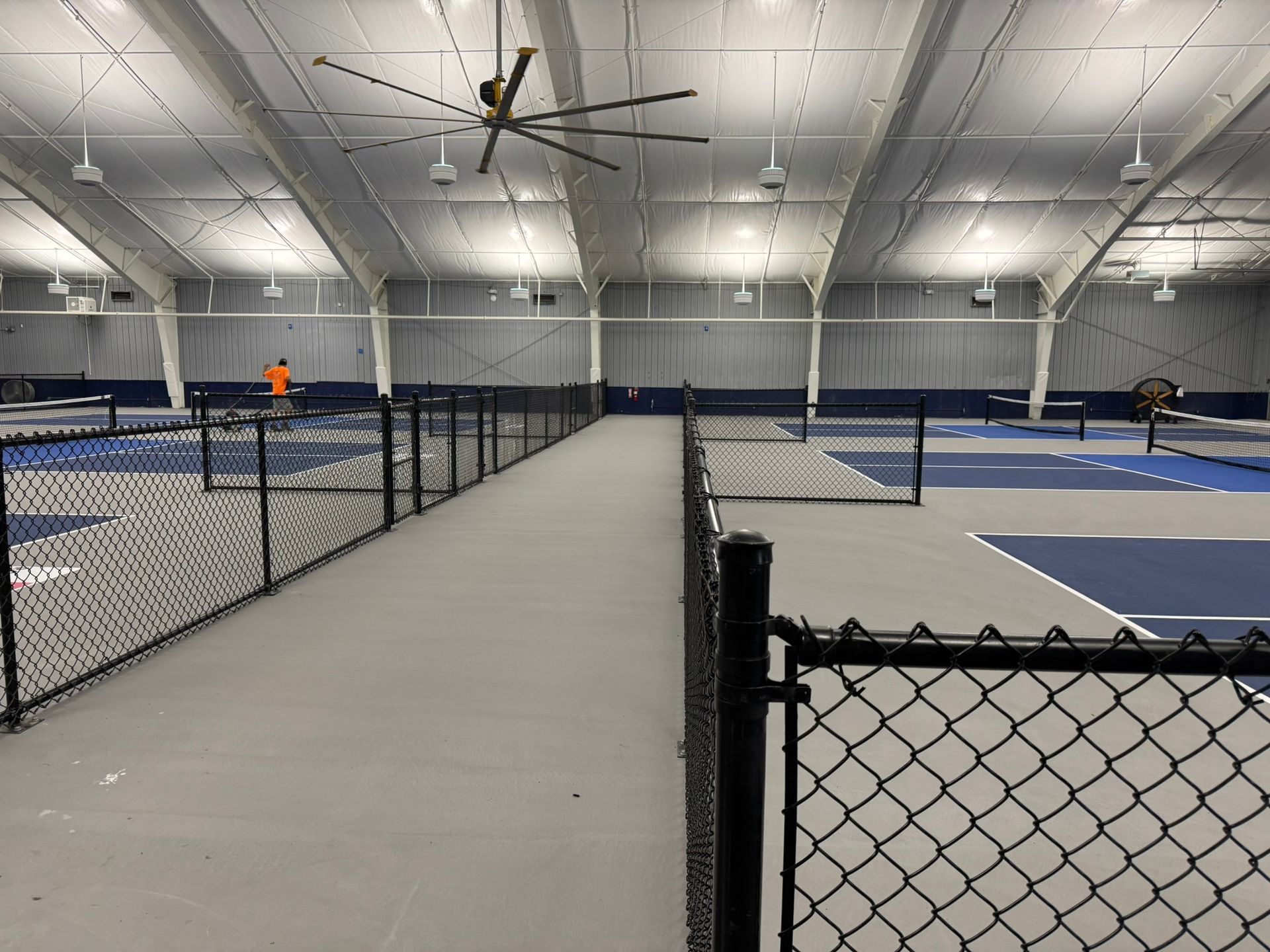 Indoor tennis court with chain-link fencing, gray walkway, and blue playing surfaces; a worker is visible in the distance.