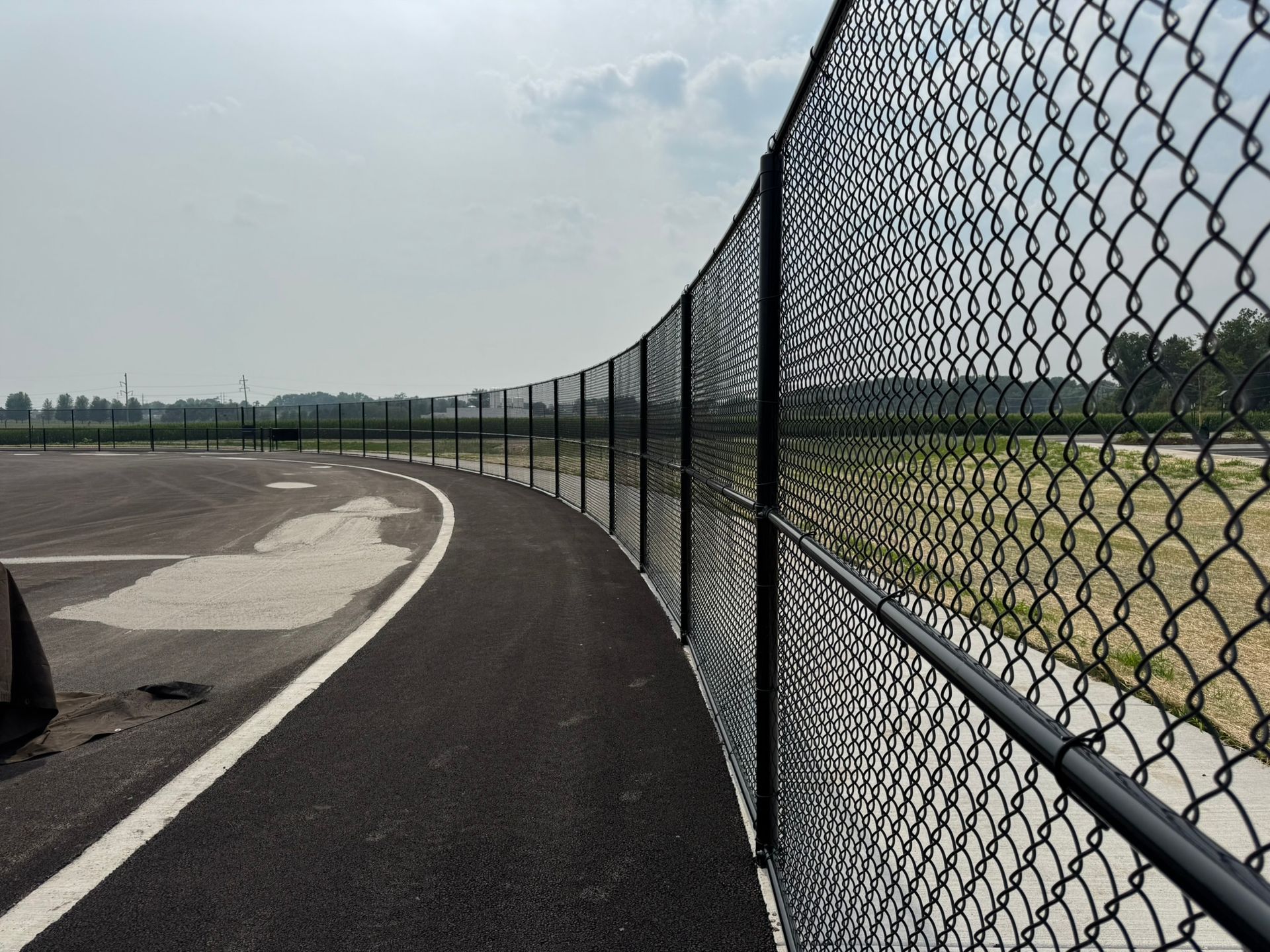 Curving black chain-link fence bordering a paved pathway; sky and distant greenery in background.
