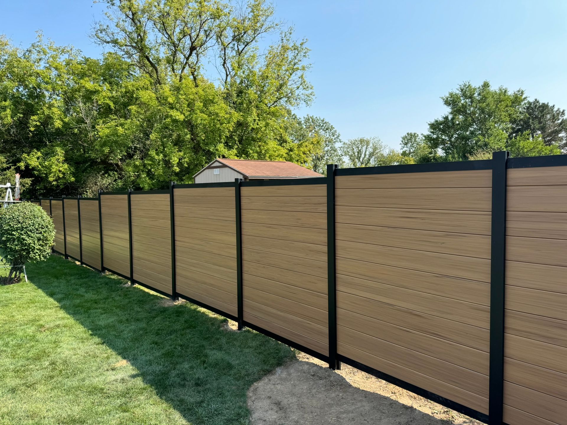Brown and black horizontal privacy fence in a grassy yard, with trees and a blue sky in the background.