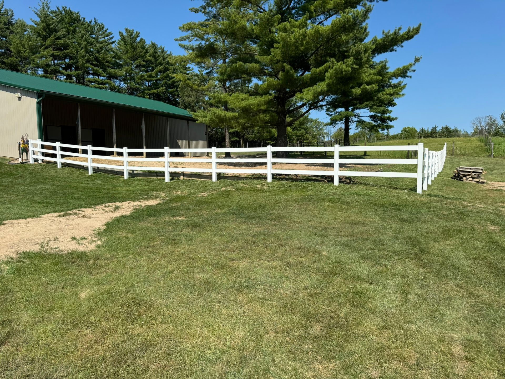 White fence surrounding a grassy area next to a building with a green roof, on a sunny day.