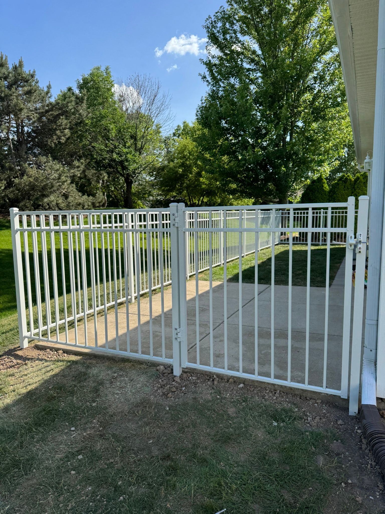 White metal fence with gate on a concrete patio, leading to a grassy area with trees and blue sky.