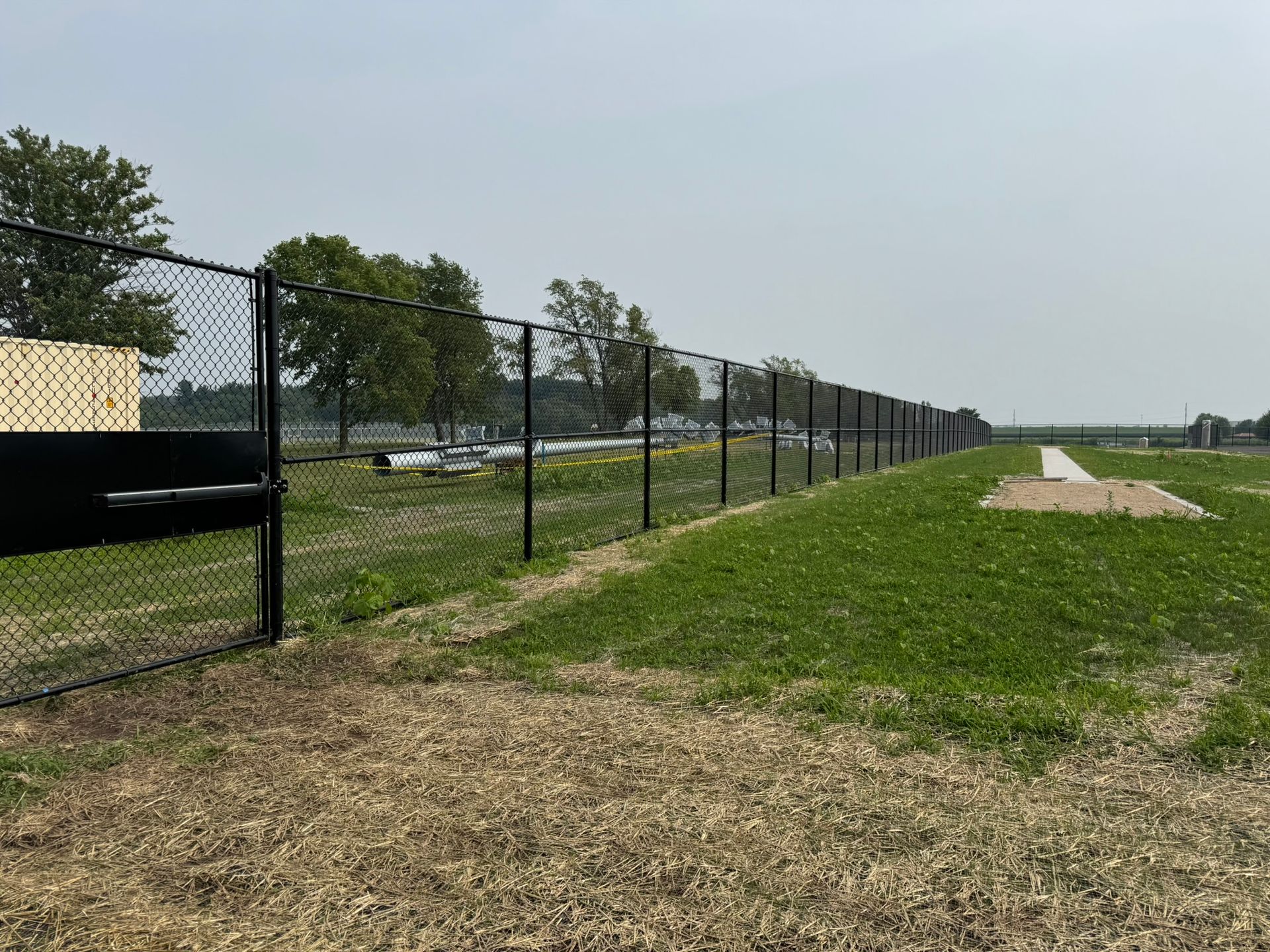 Black chain-link fence bordering a grassy field, under a hazy sky.