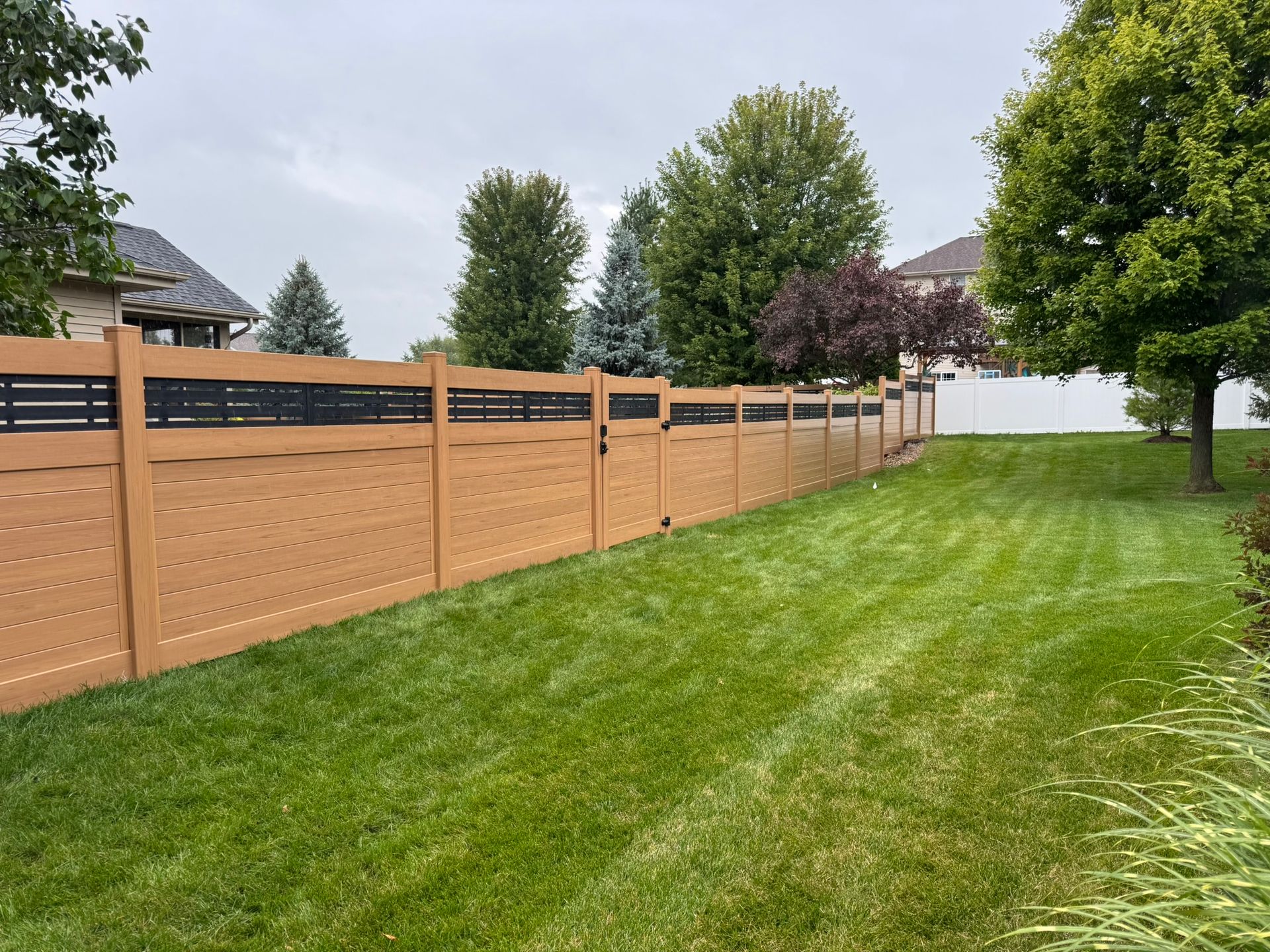 Brown and black fenced backyard with green grass and trees.