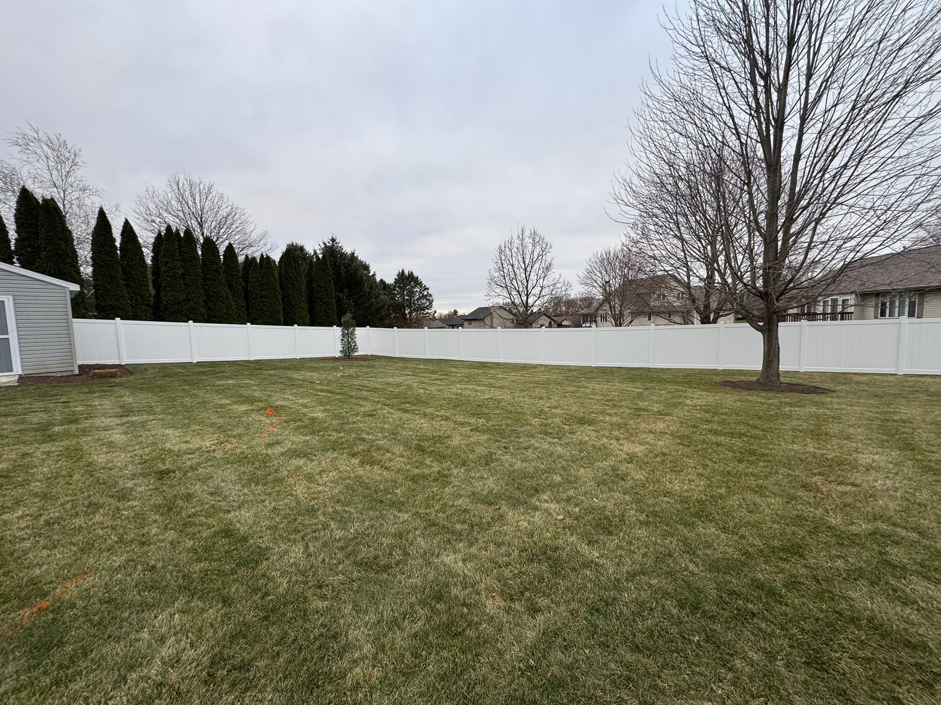 Backyard with white fence, green grass, and trees under a cloudy sky.