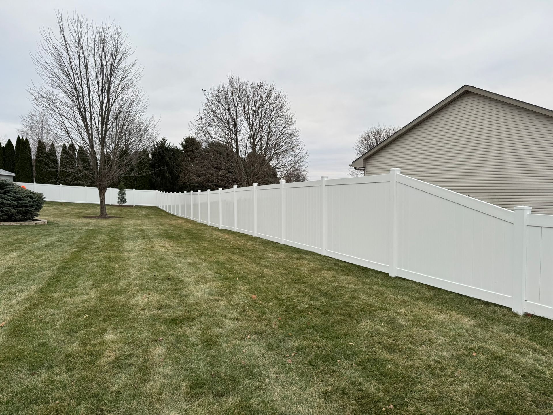 White vinyl fence along a grassy backyard, cloudy sky, bare trees, and a house.