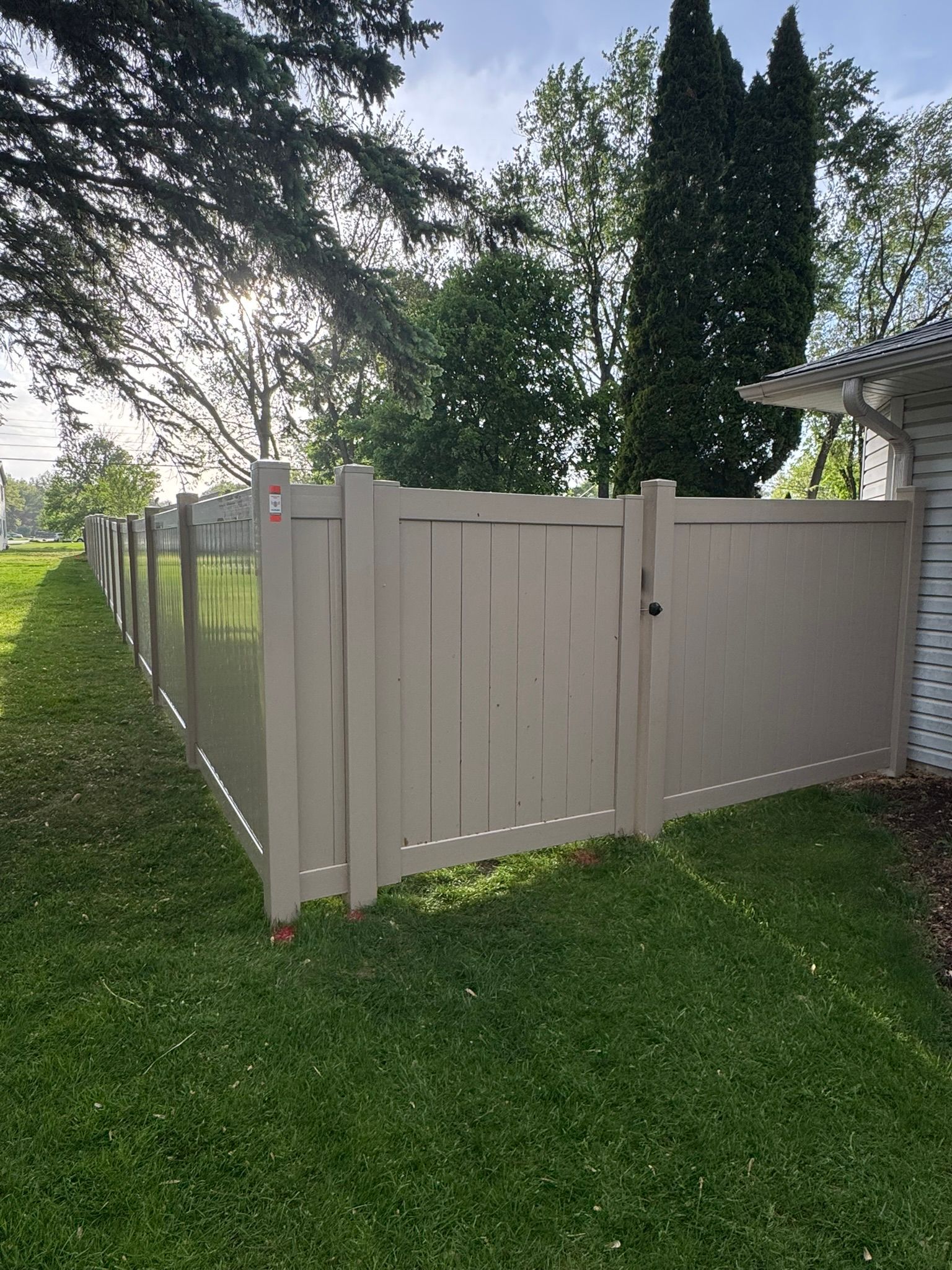Tan vinyl fence with gate, bordering green lawn, trees, and partial white building.