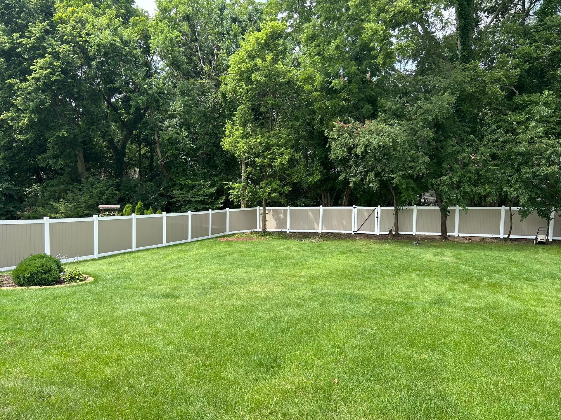 A fenced backyard with green grass and trees. The fence is tan and white.