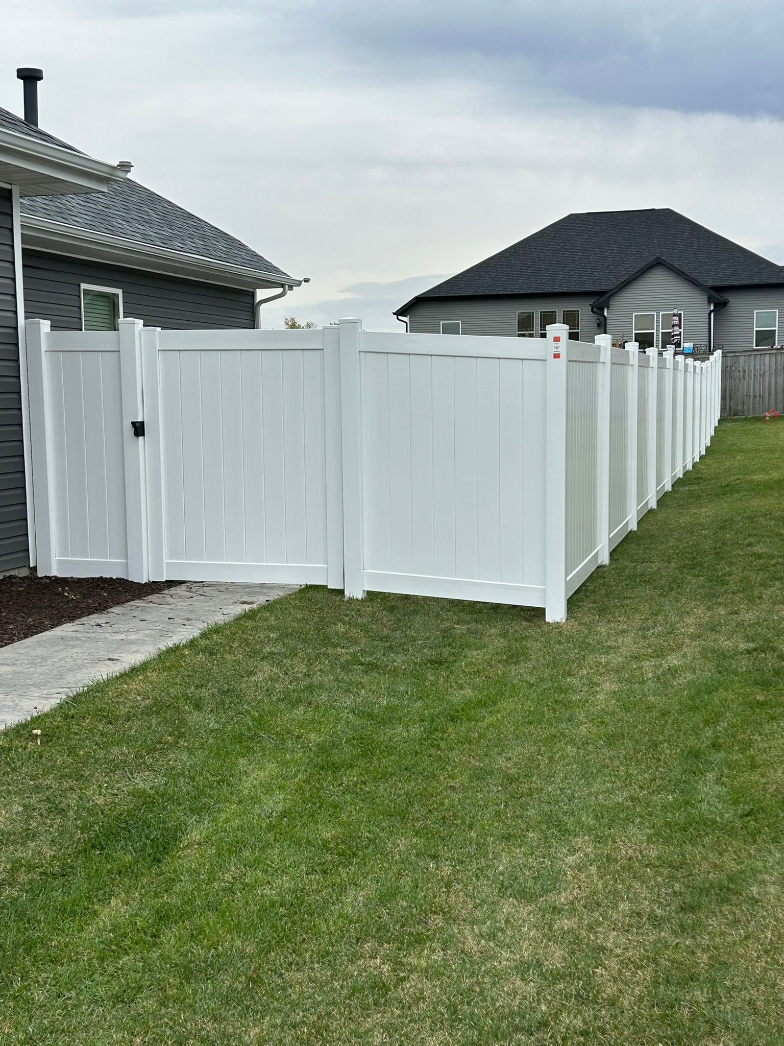 White vinyl fence enclosing a green lawn between two houses. Cloudy sky in background.