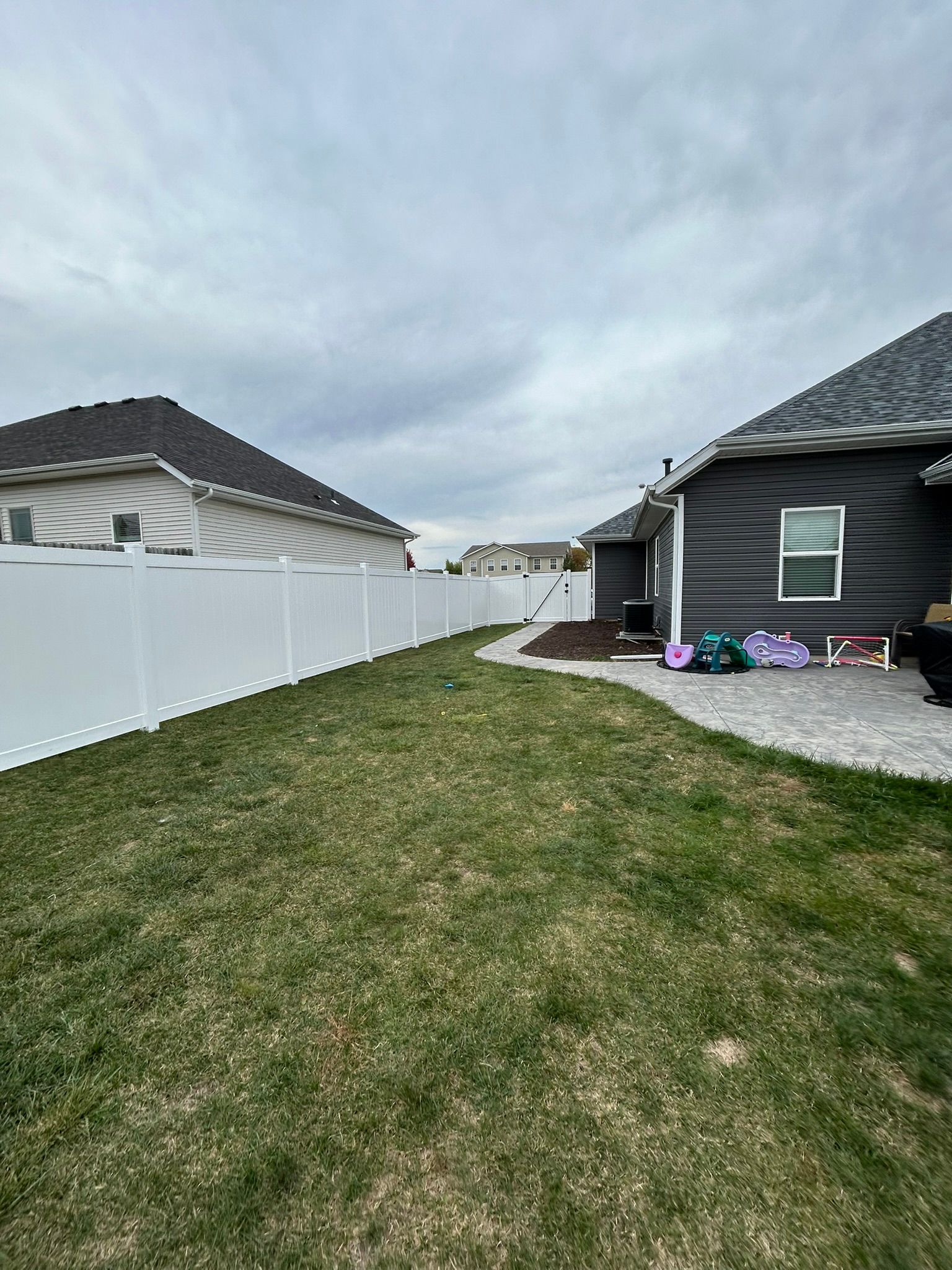 Lawn in a backyard with a white fence, gray house, and cloudy sky.