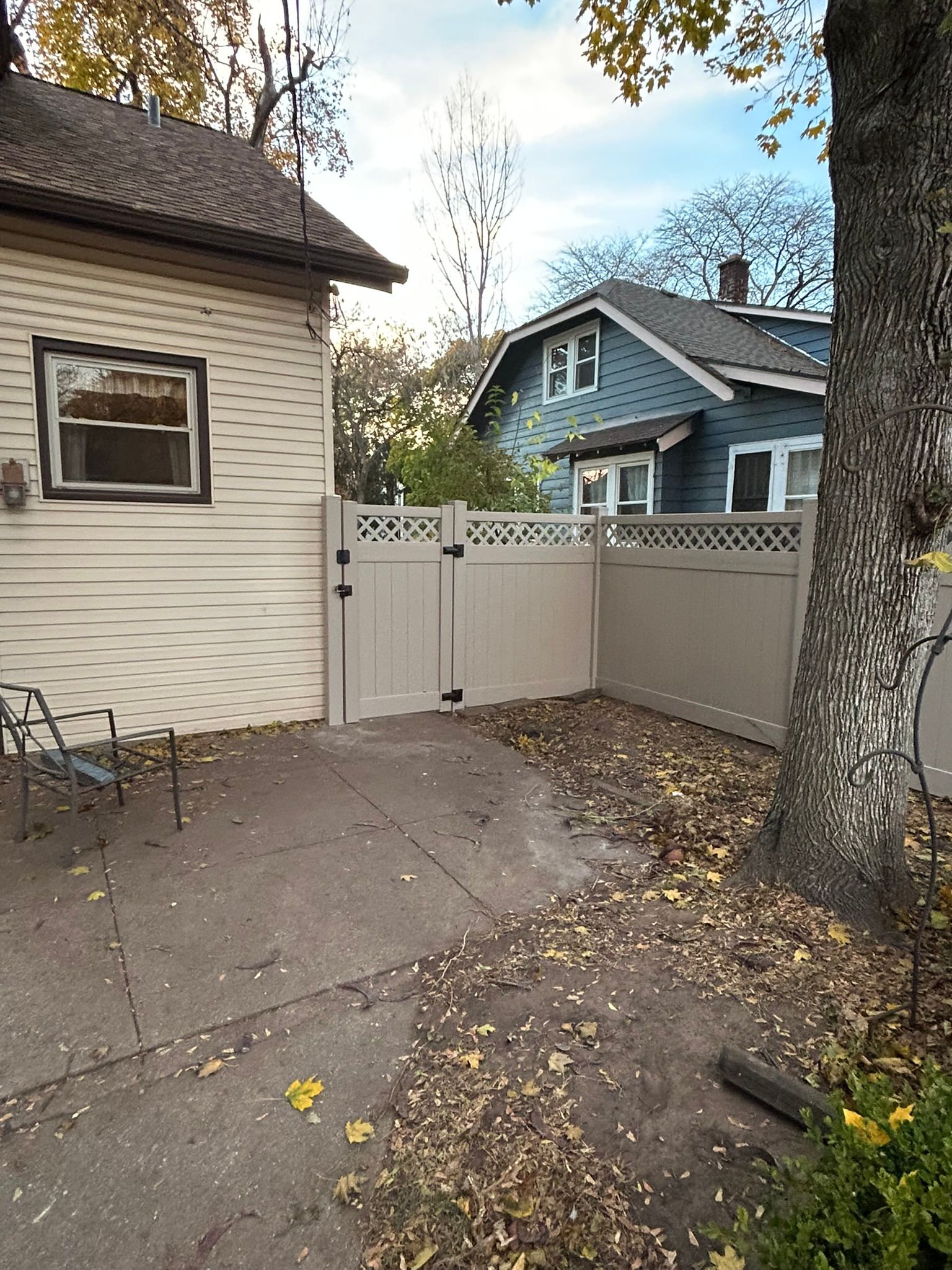 Backyard with tan fence, gate, concrete patio, a house, and a tree with fallen leaves.