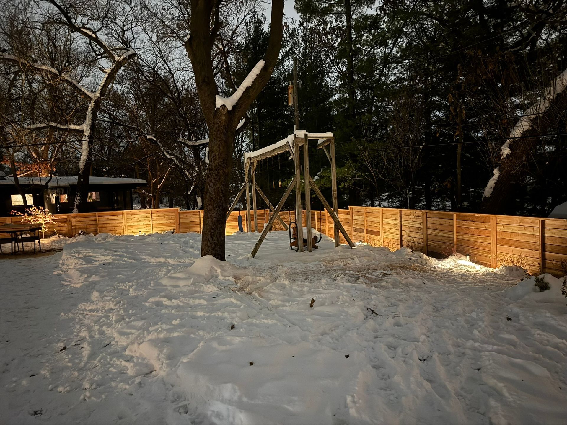 Snowy backyard at dusk with wooden swing set and fence. Lit by warm lights. Tree in center.