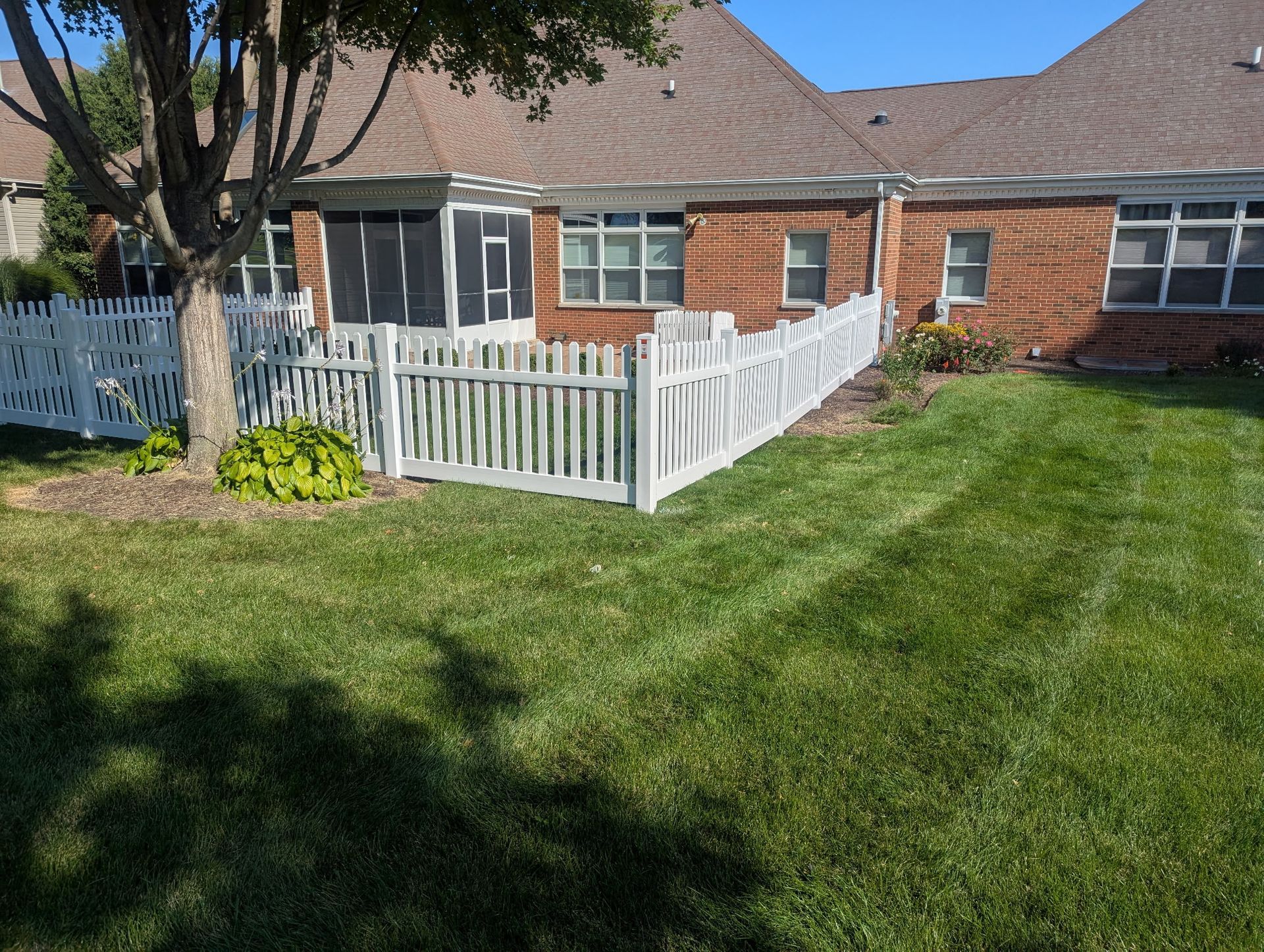 White picket fence surrounds a backyard with green grass and a brick house.