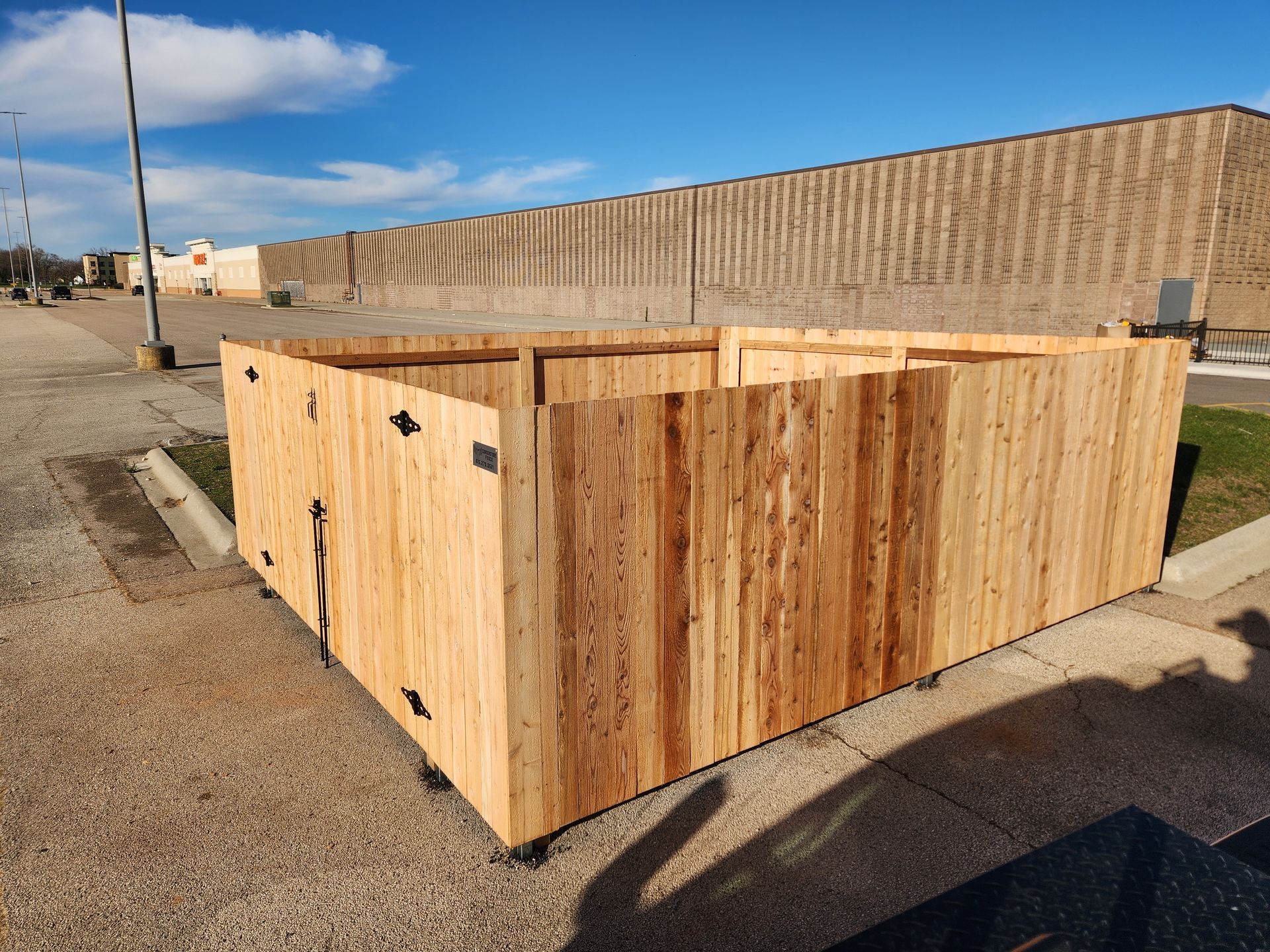 Wooden structure, possibly a maze, sits on asphalt parking lot, next to a large building under a blue sky.