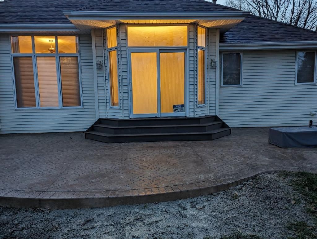 Back of house at dusk, lit windows, stone patio, steps leading up, grey siding, outdoor setting.