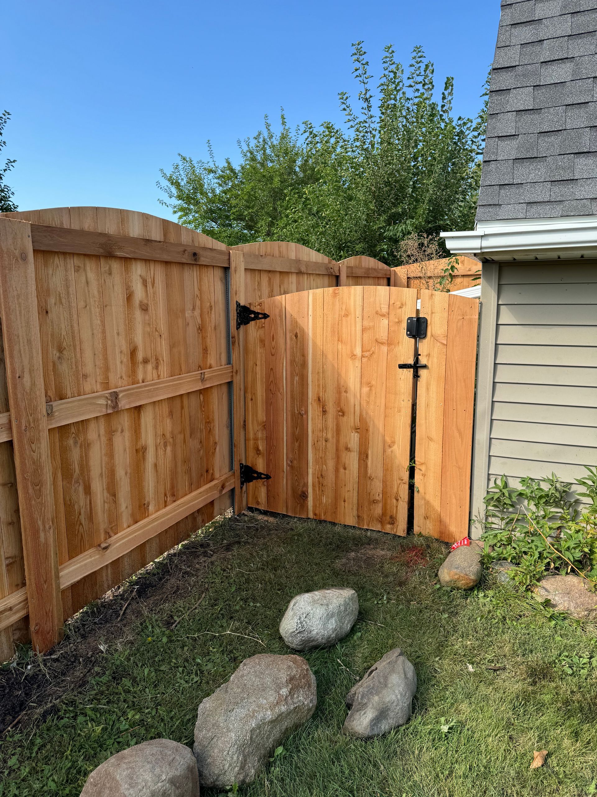 Wooden fence with a gate, rocks on the ground, and a chicken near a building with a light-colored exterior.