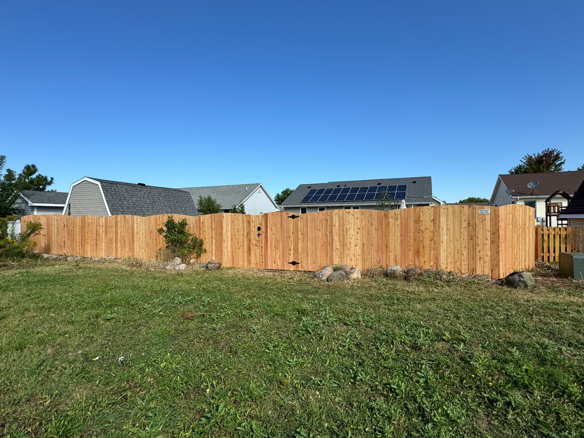 Wooden fence in front of green lawn with houses and blue sky in the background.