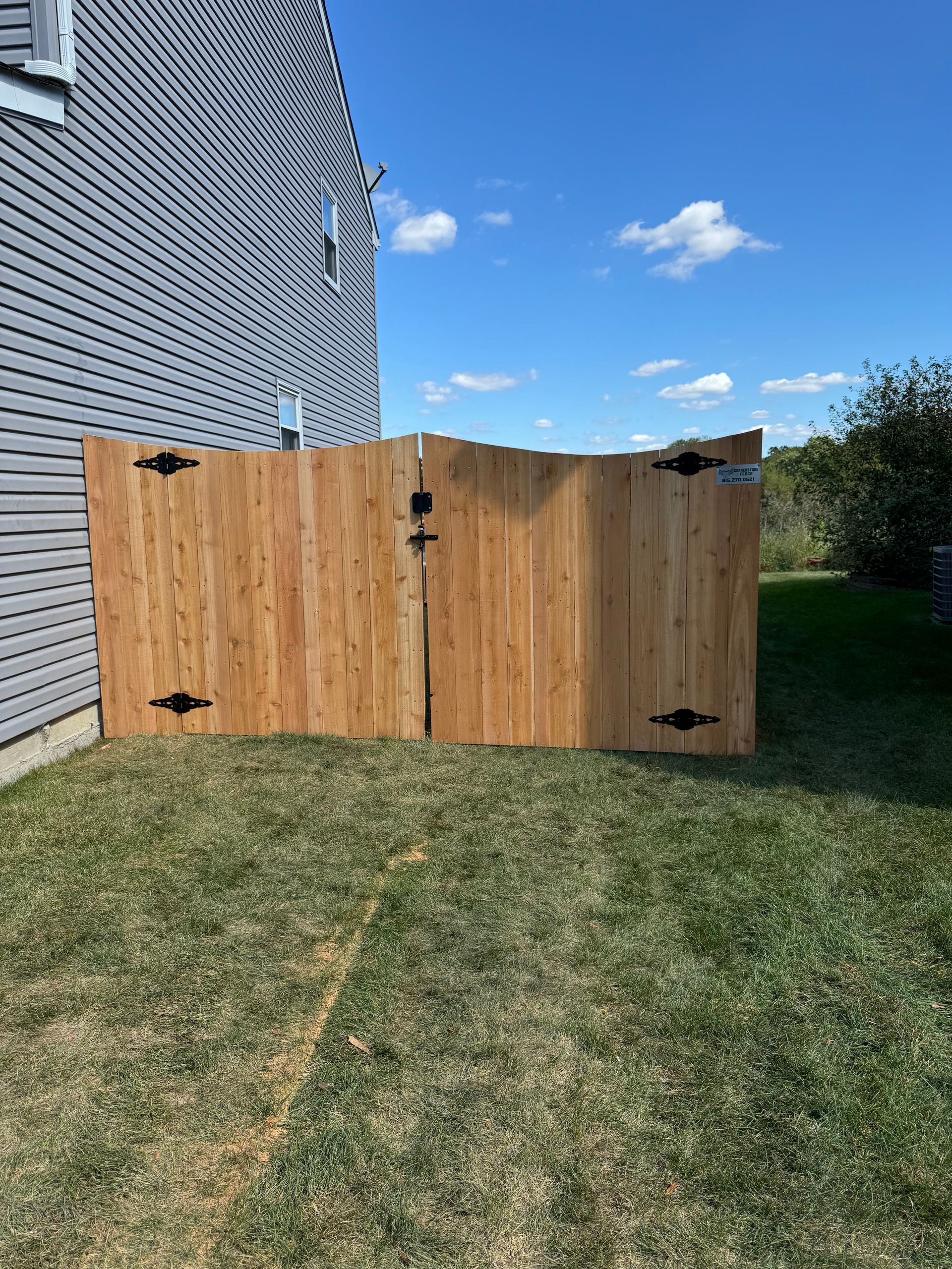 Two wooden fence sections with black hinges on a grassy lawn next to a gray house under a blue sky.