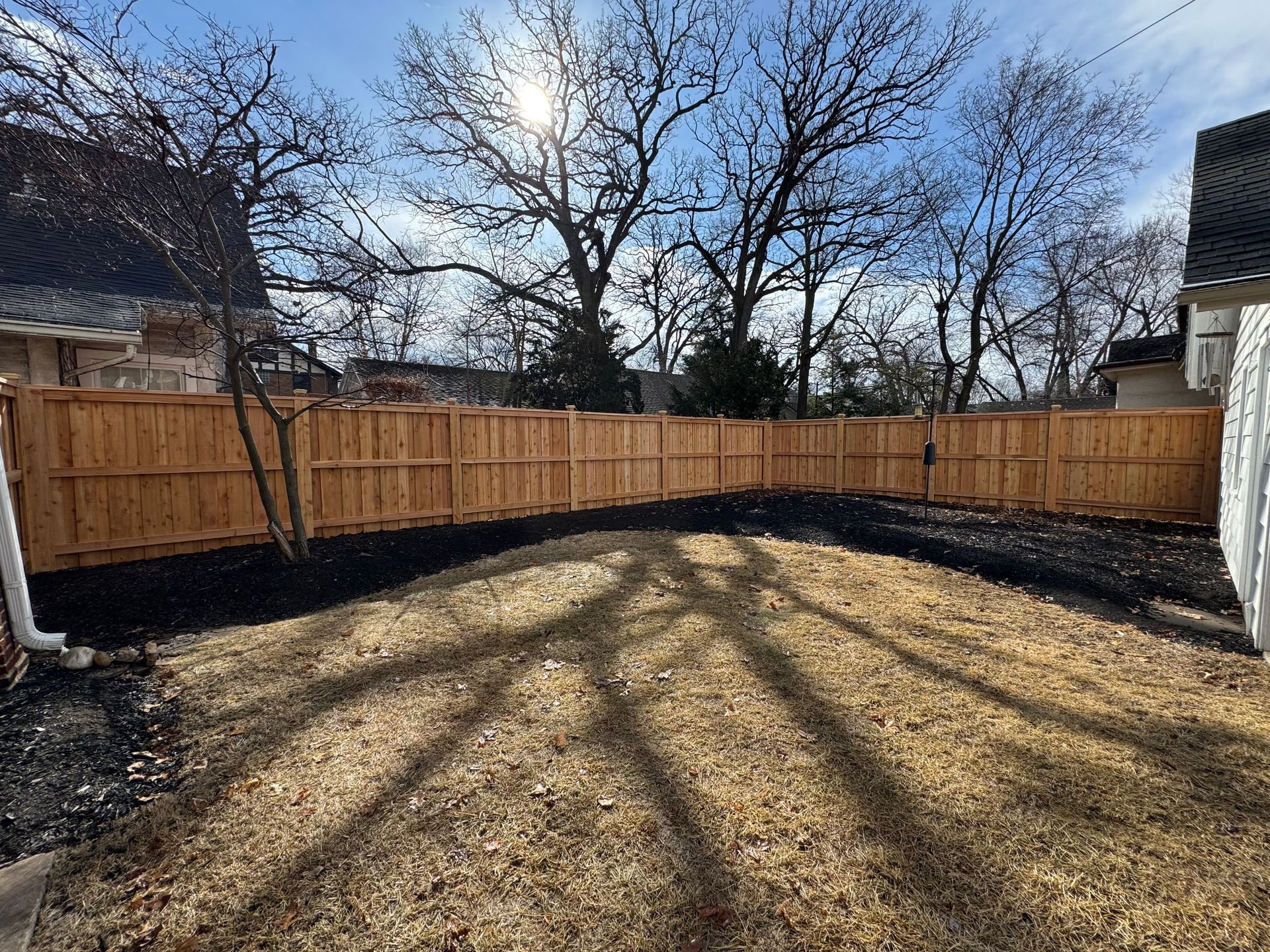 Wooden fence surrounds a backyard with shadows from trees, under a sunny sky.