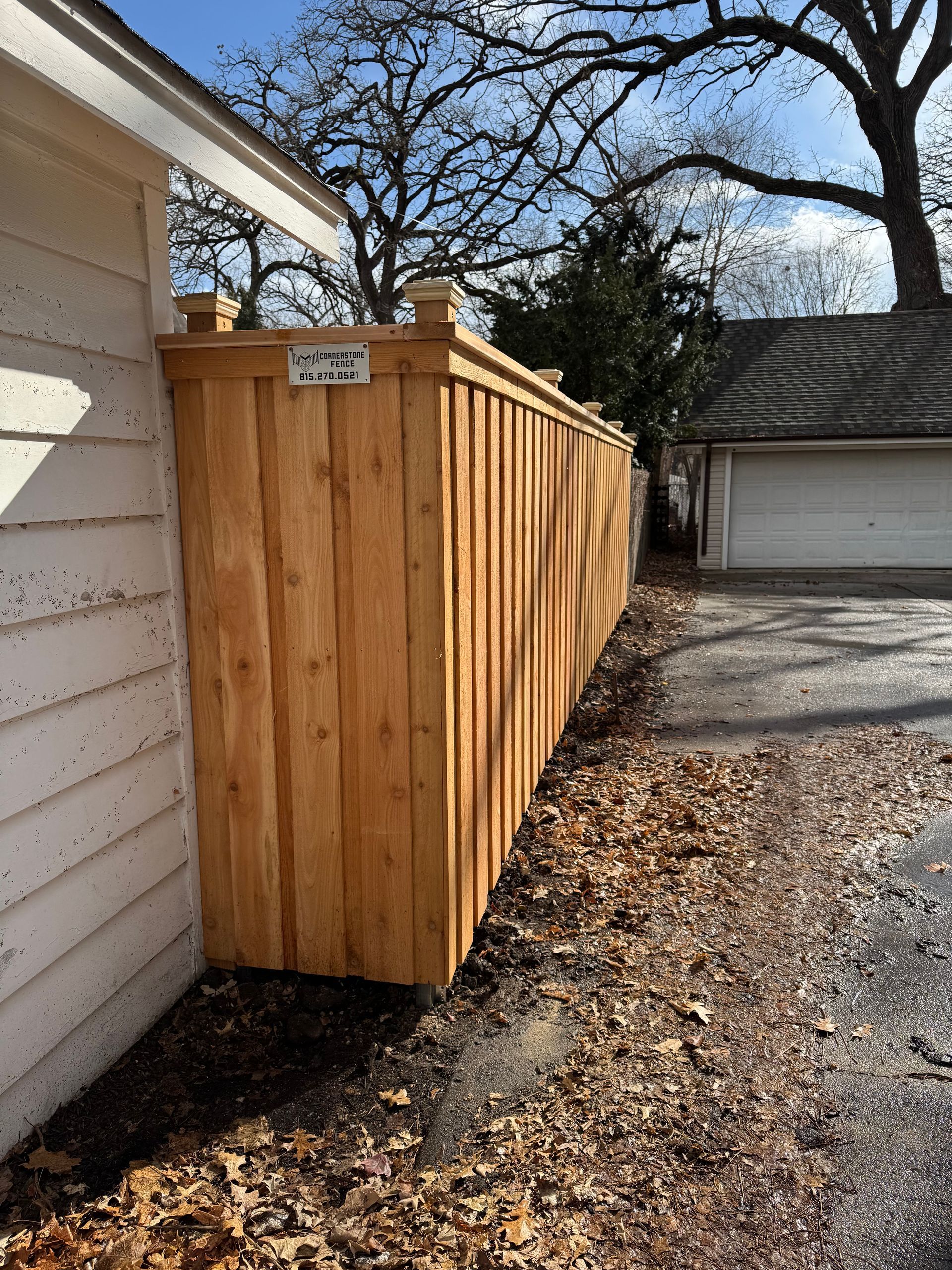 Wooden fence alongside a building and driveway, with a leaflittered ground. Sunny day.
