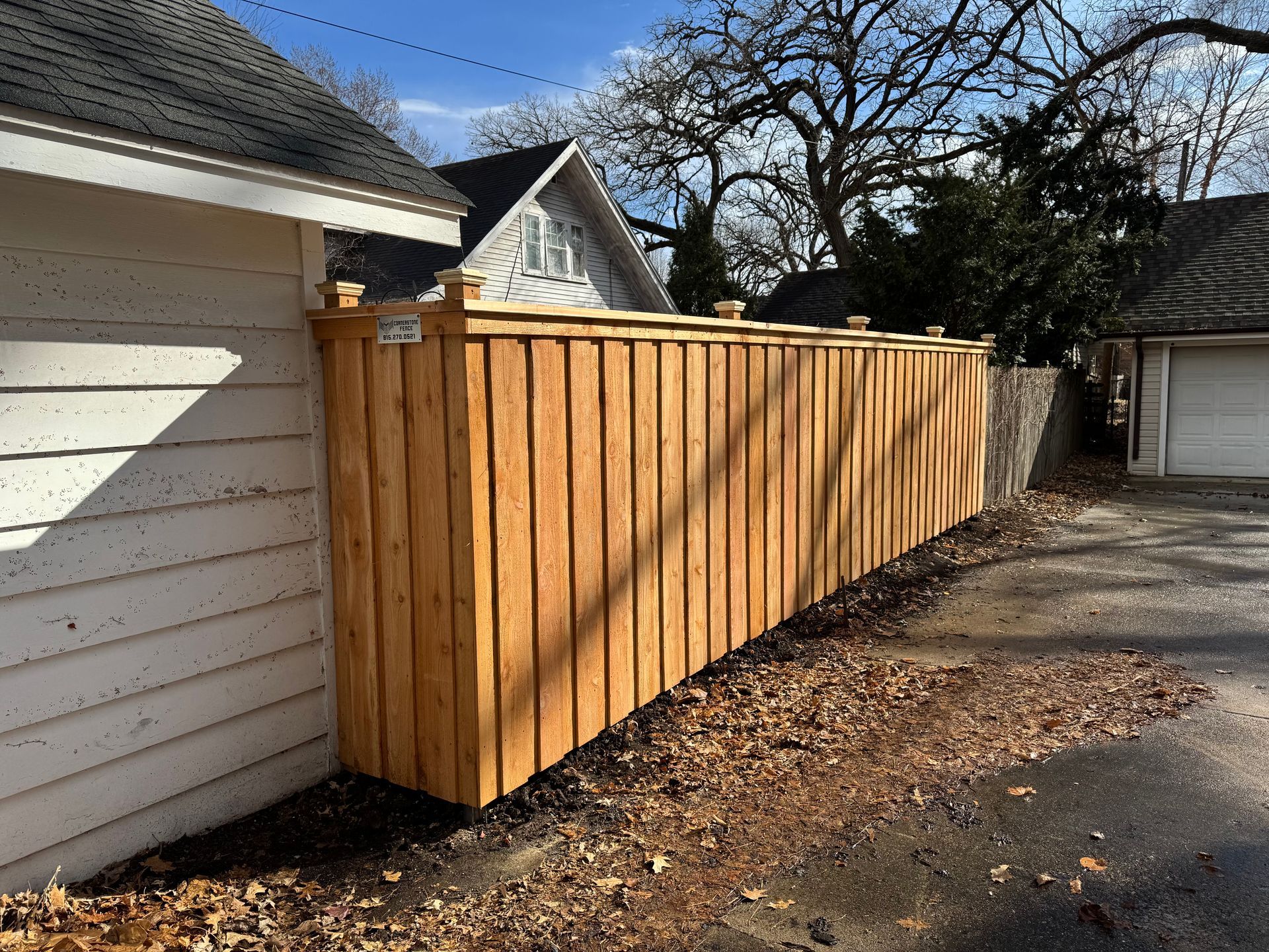 Wooden fence along a driveway, next to a white building. Autumn leaves on the ground, sunny day.