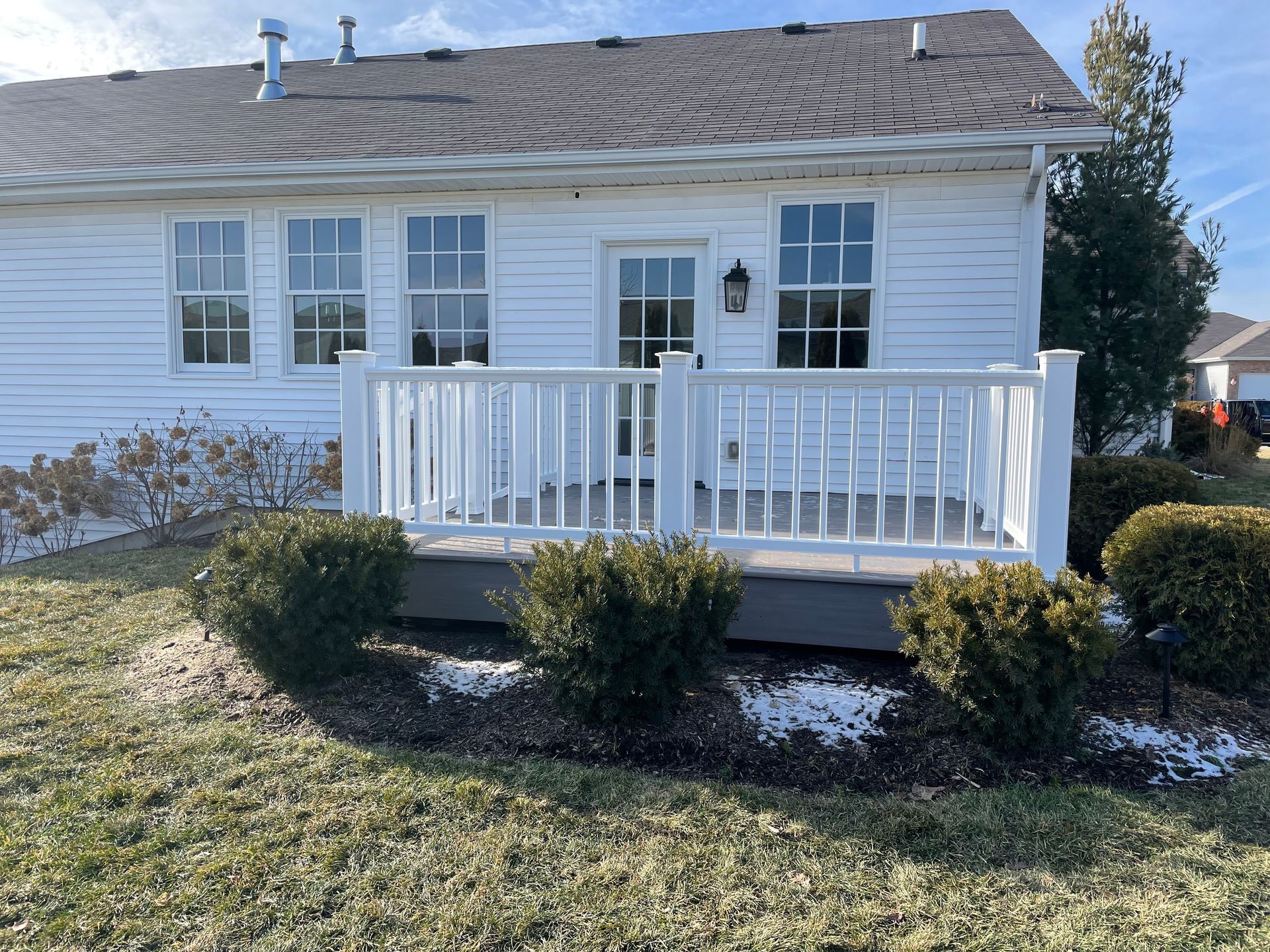 White house exterior with deck and bushes; brown roof, green grass, and blue sky.