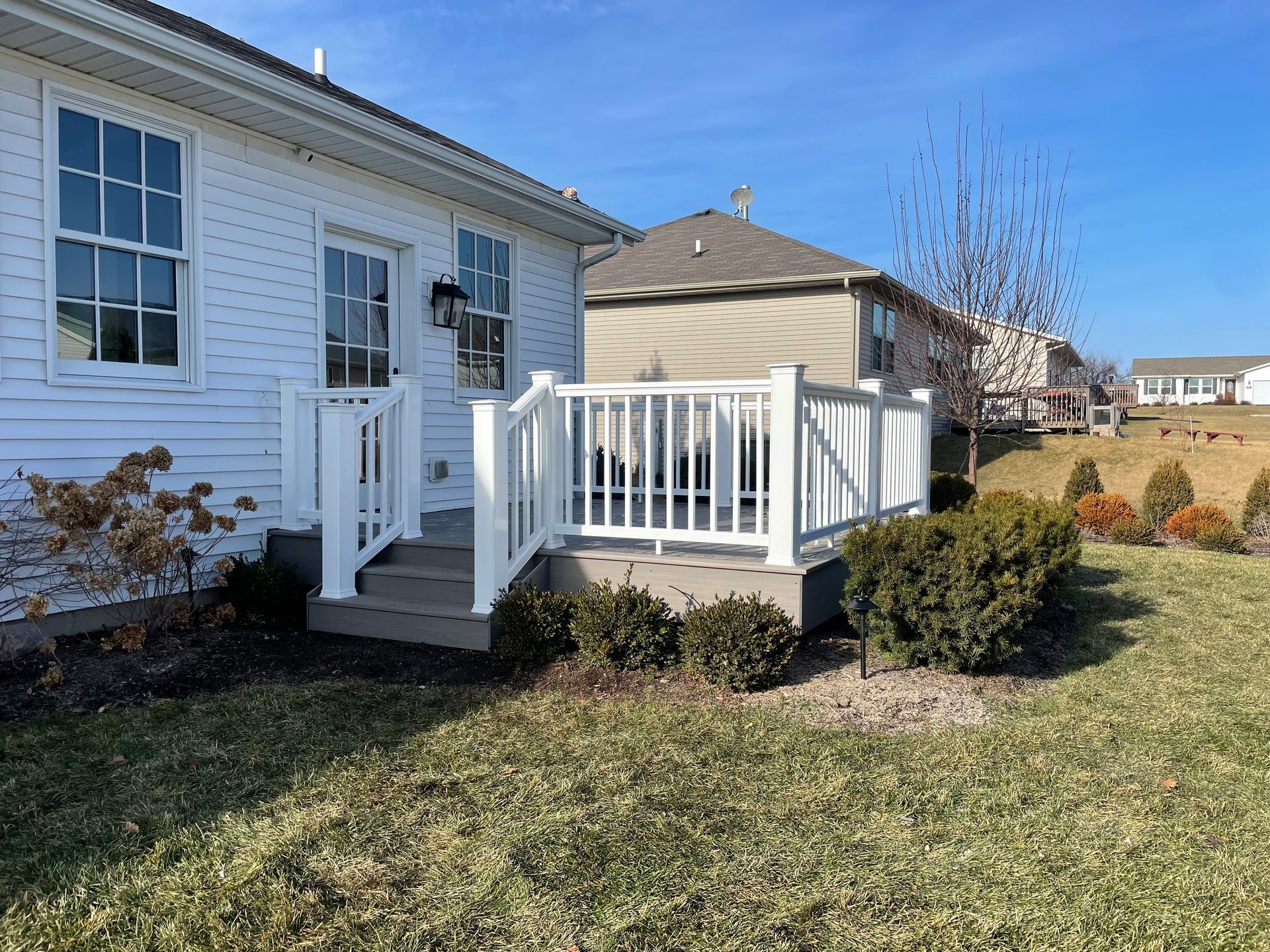 White house with a small deck and railing, overlooking a grassy yard on a sunny day.