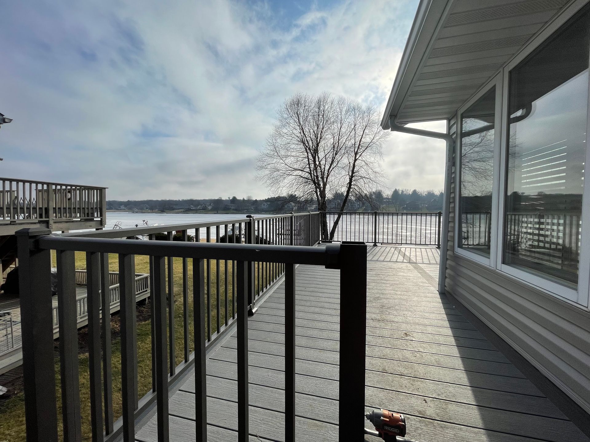 Deck overlooking a lake on a cloudy day. Grey railings and house siding; tree in the distance.