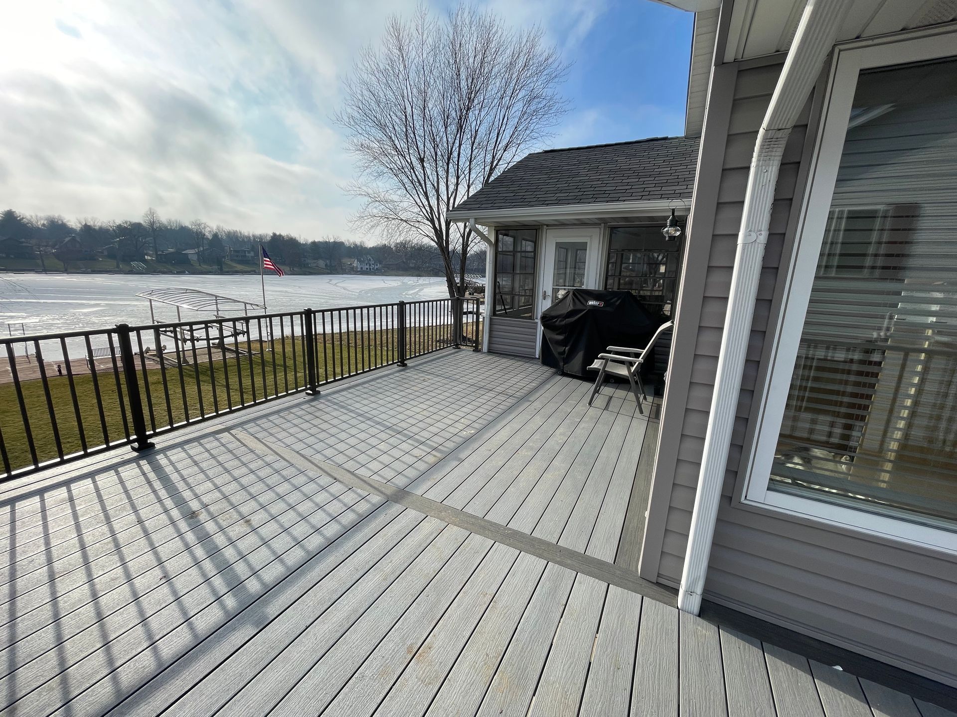Gray deck overlooking a frozen lake, next to a house with a door, grill, and window.