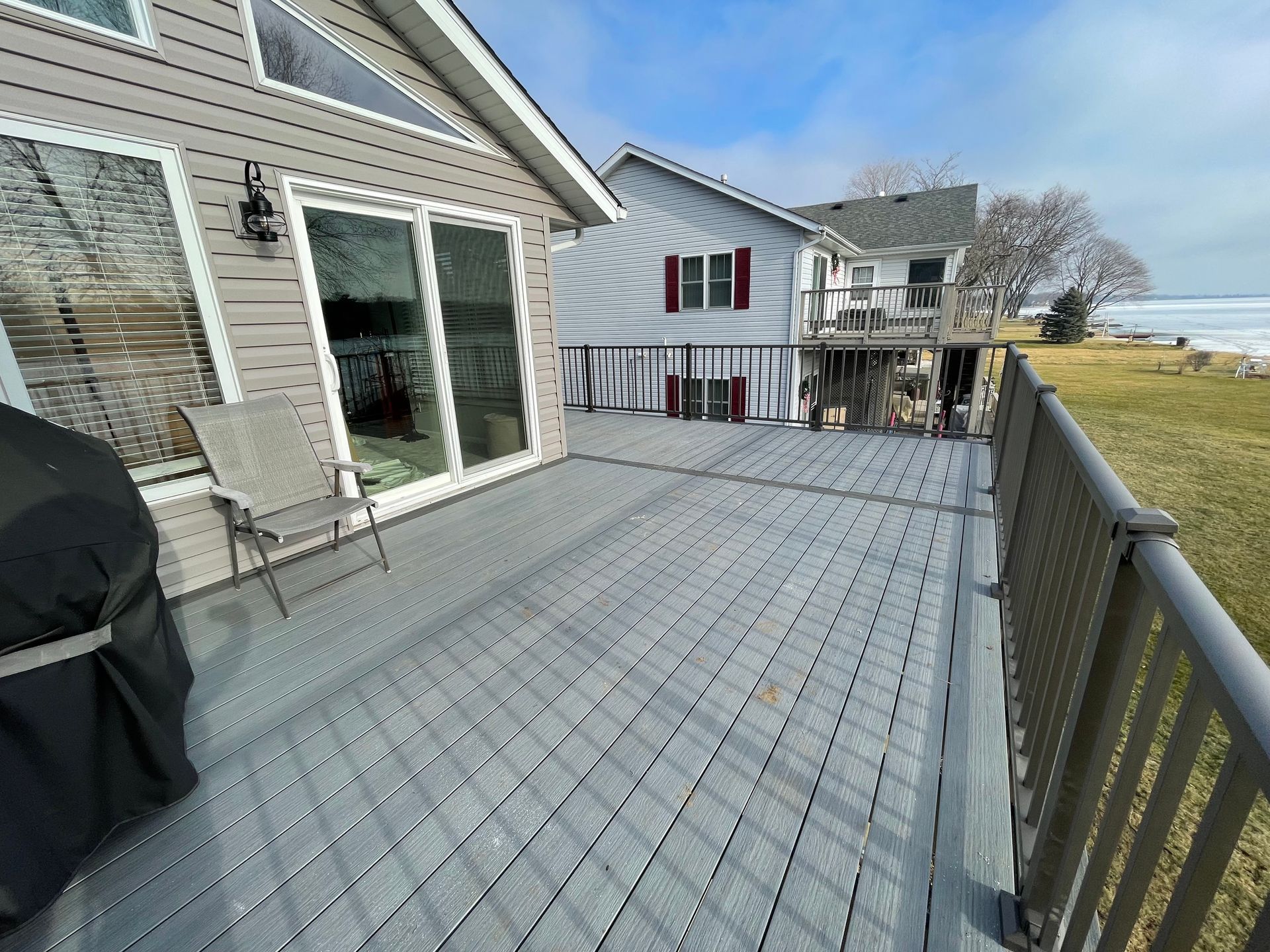 Grey wooden deck alongside a house, with a lake and blue sky visible.