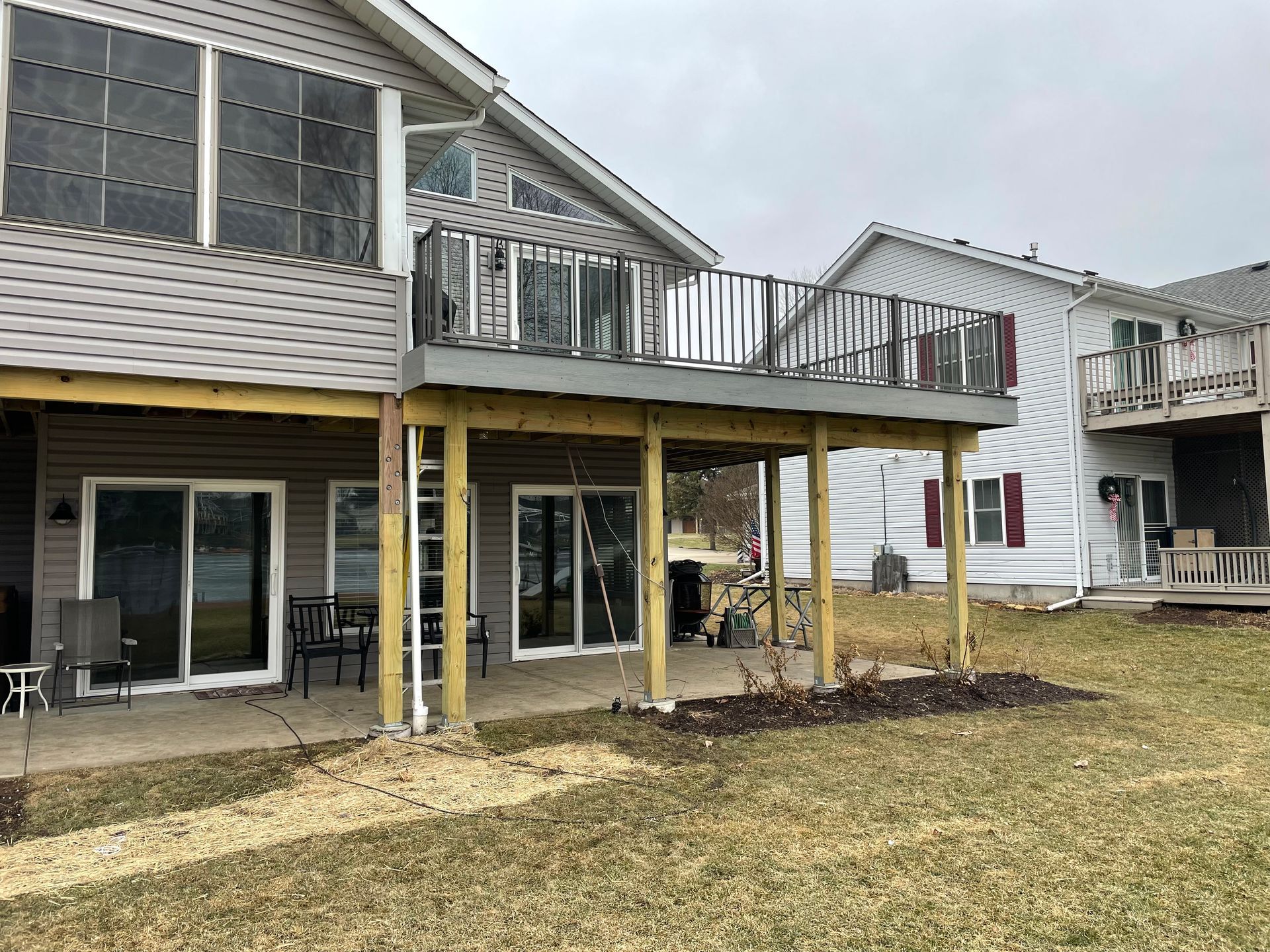 Deck extending from a multi-story house with sliding doors. Supported by wooden posts, surrounded by grass.