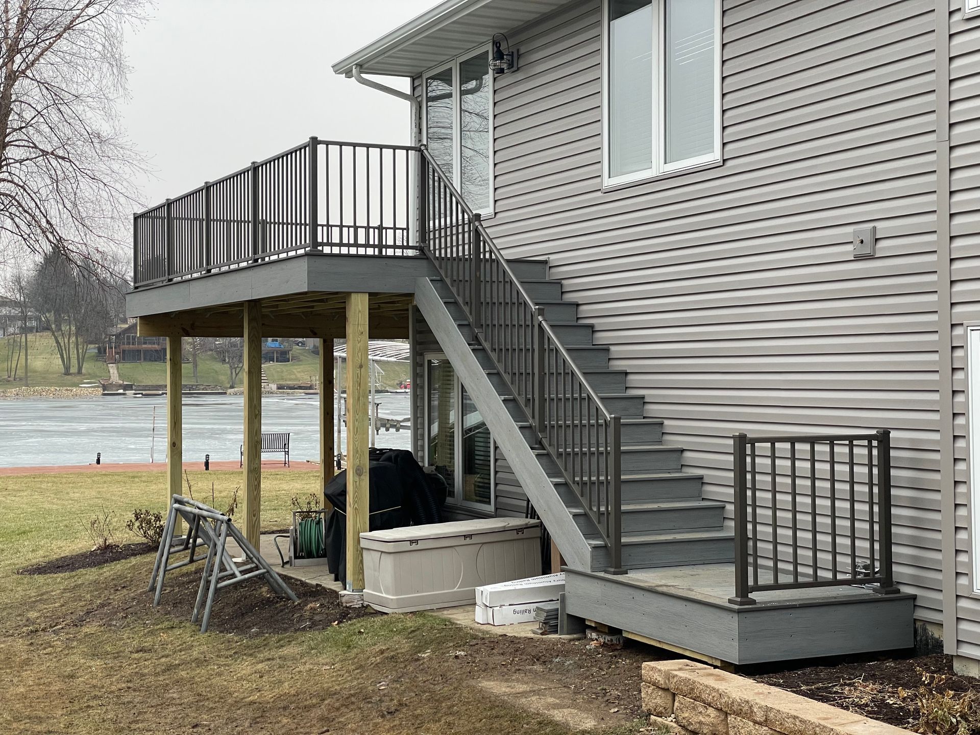 Deck with stairs, gray siding house, black railings.  Lake view in the background.