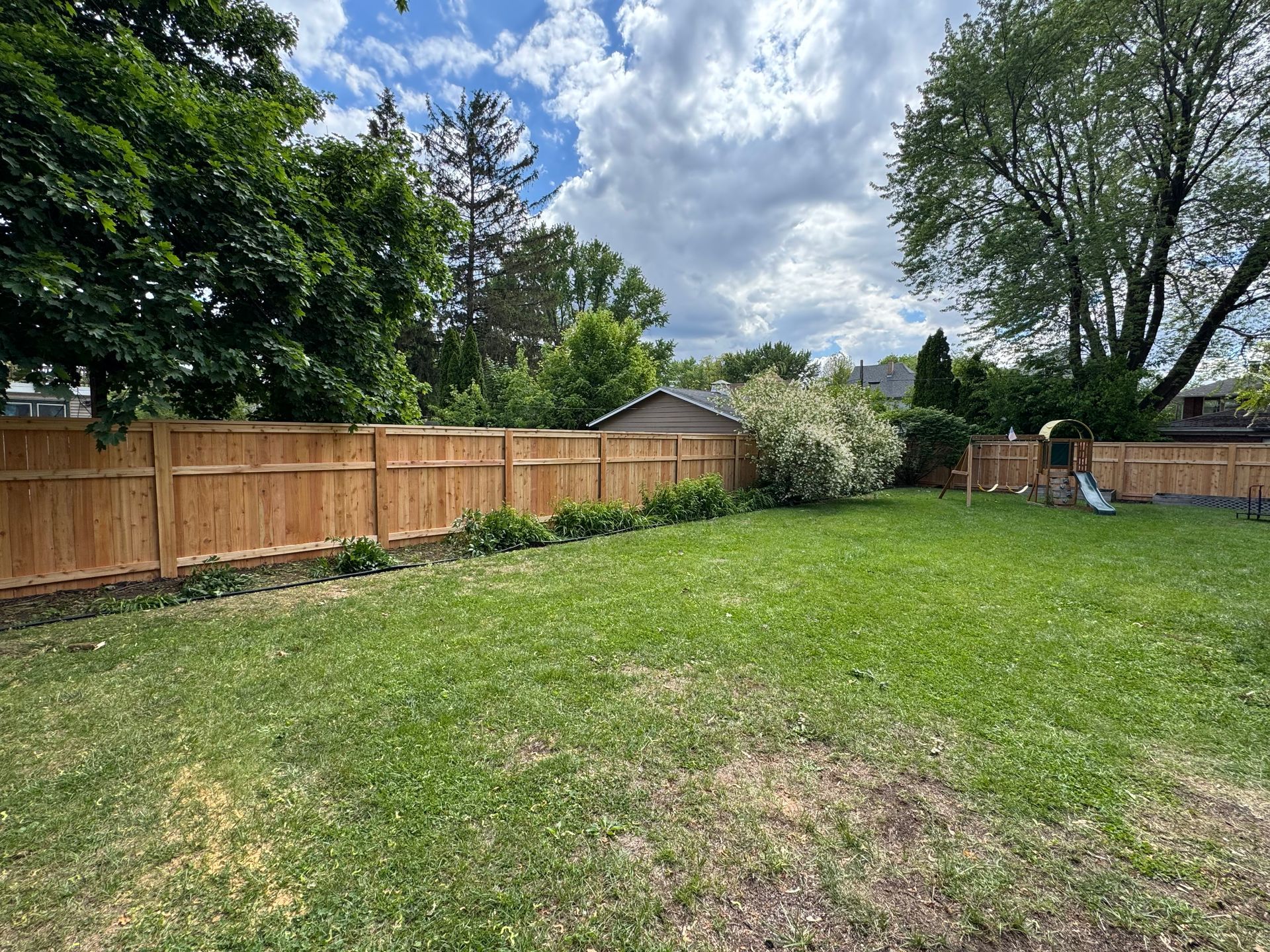 A wooden fence encloses a green backyard with grass, trees, and a partly cloudy sky.