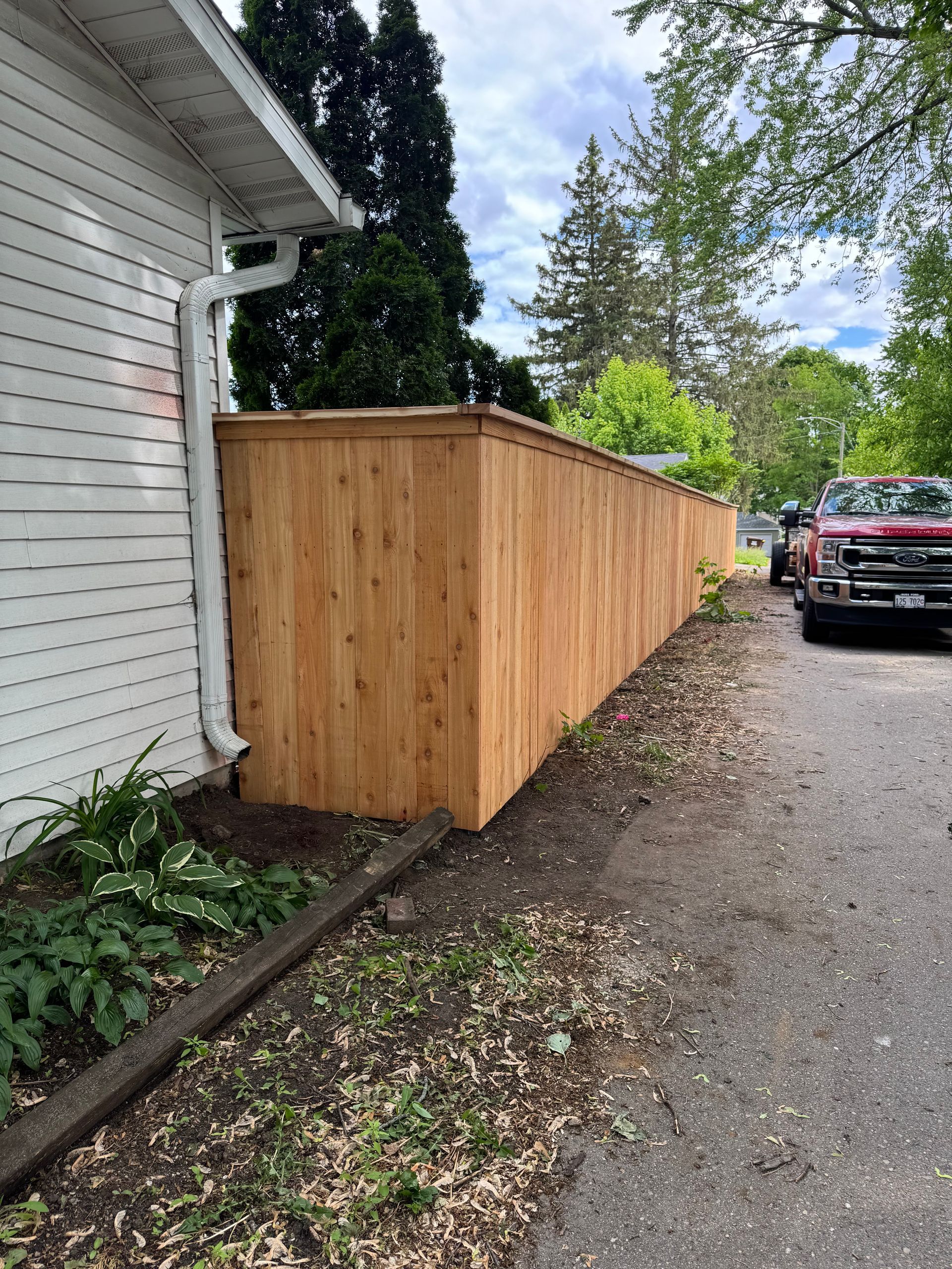 Wooden fence bordering a white house and a driveway, under a cloudy sky.