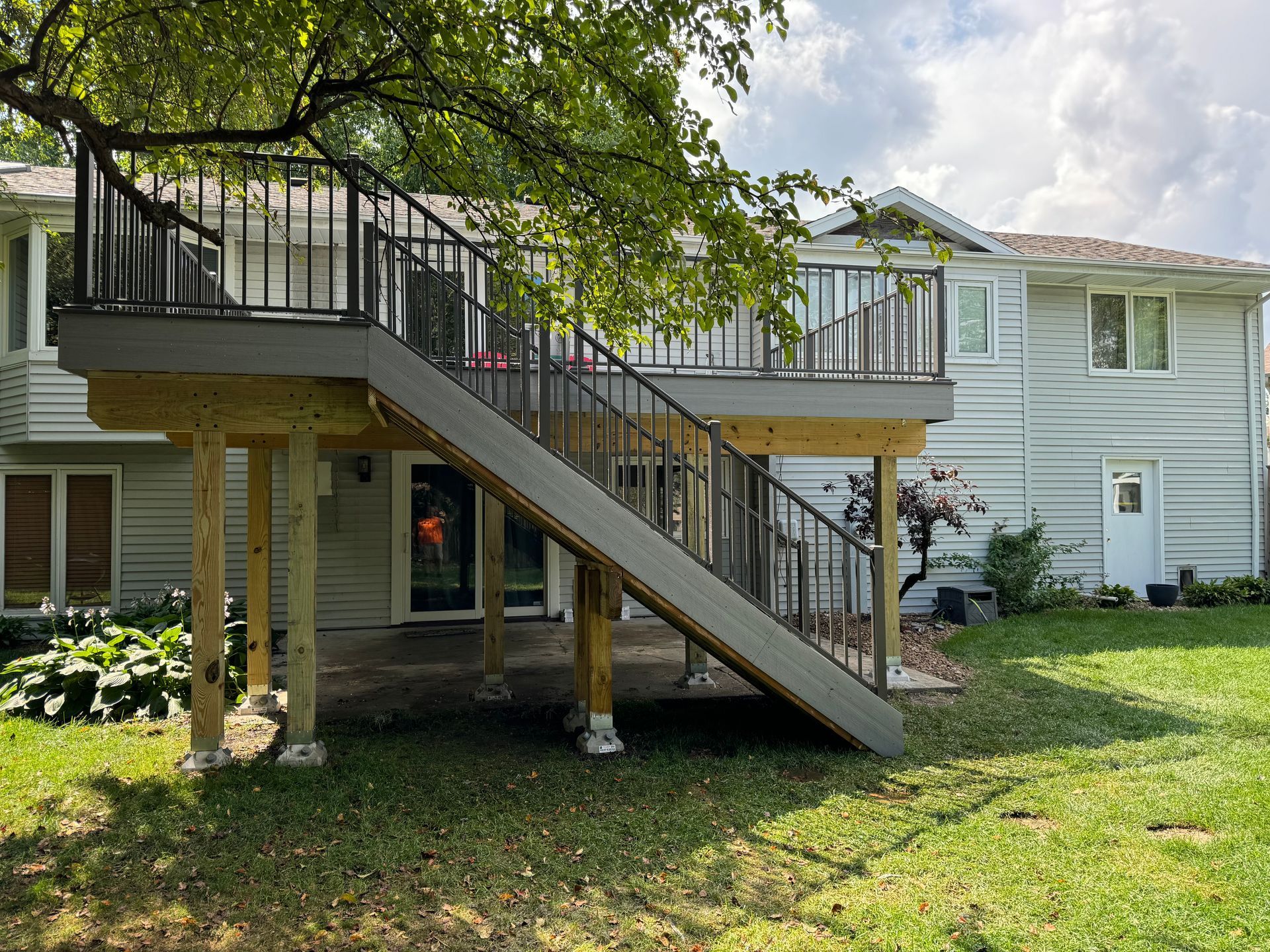 A two-story deck with stairs connected to a light-colored house. Green grass and trees surround it.