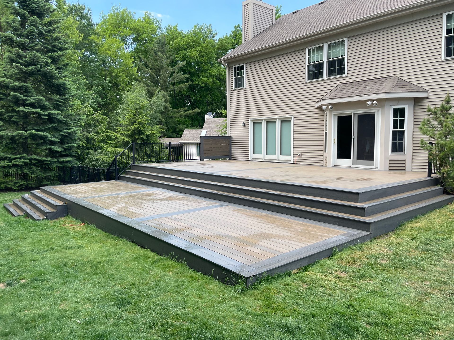 Backyard patio with steps, gray deck, and a two-story beige house. Green grass and trees surround it.