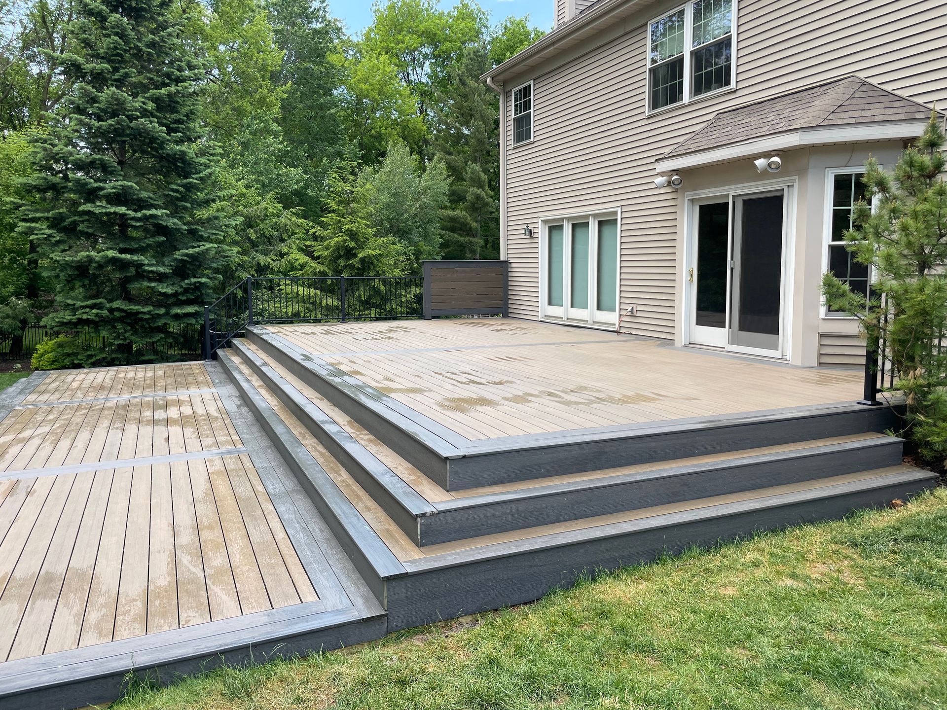 Backyard deck with steps leading to a house, surrounded by grass and trees. Gray deck with light brown accents.