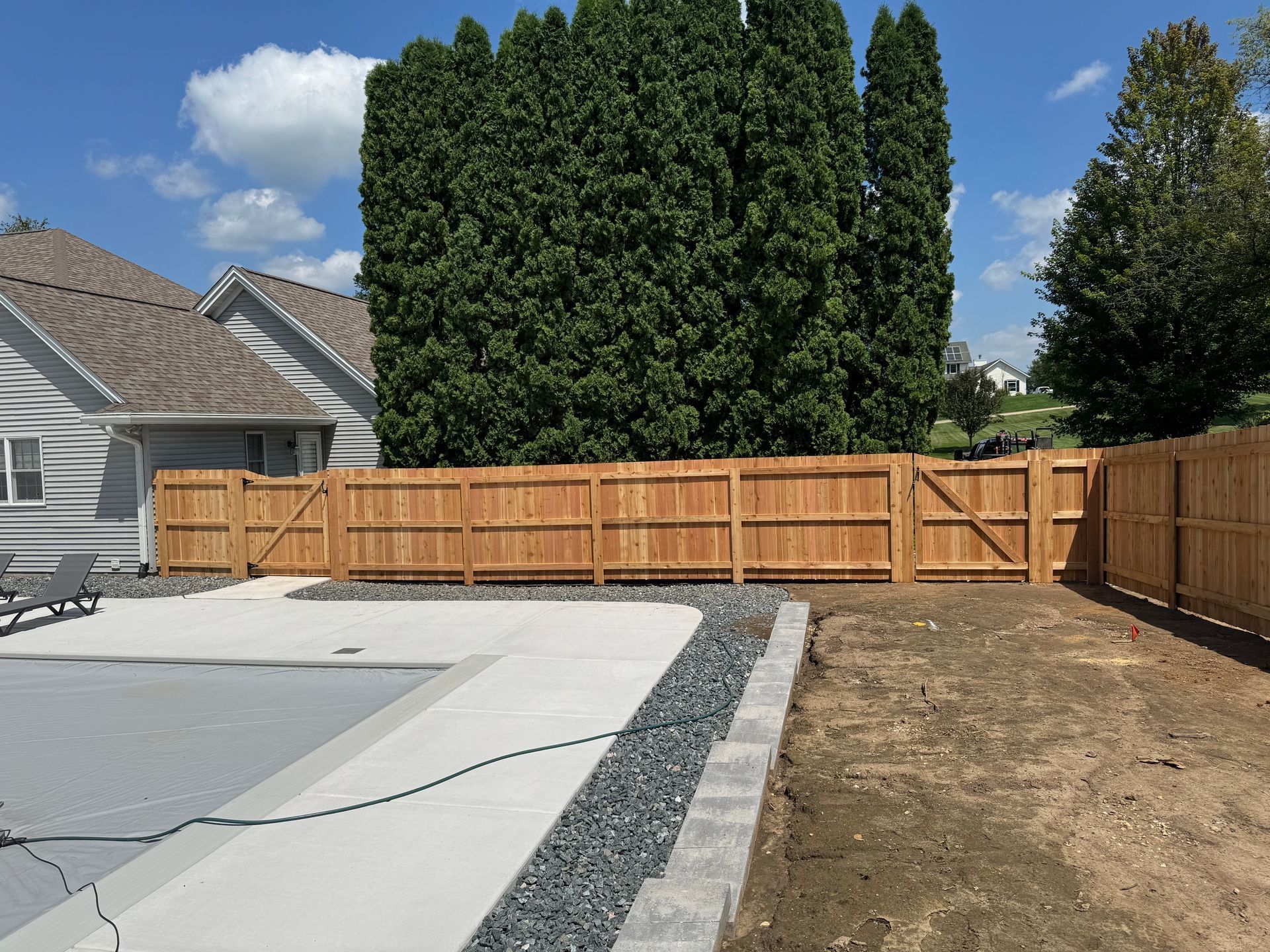 Wooden fence surrounding a backyard with a pool. Cedar fence with two gates, large green trees behind.