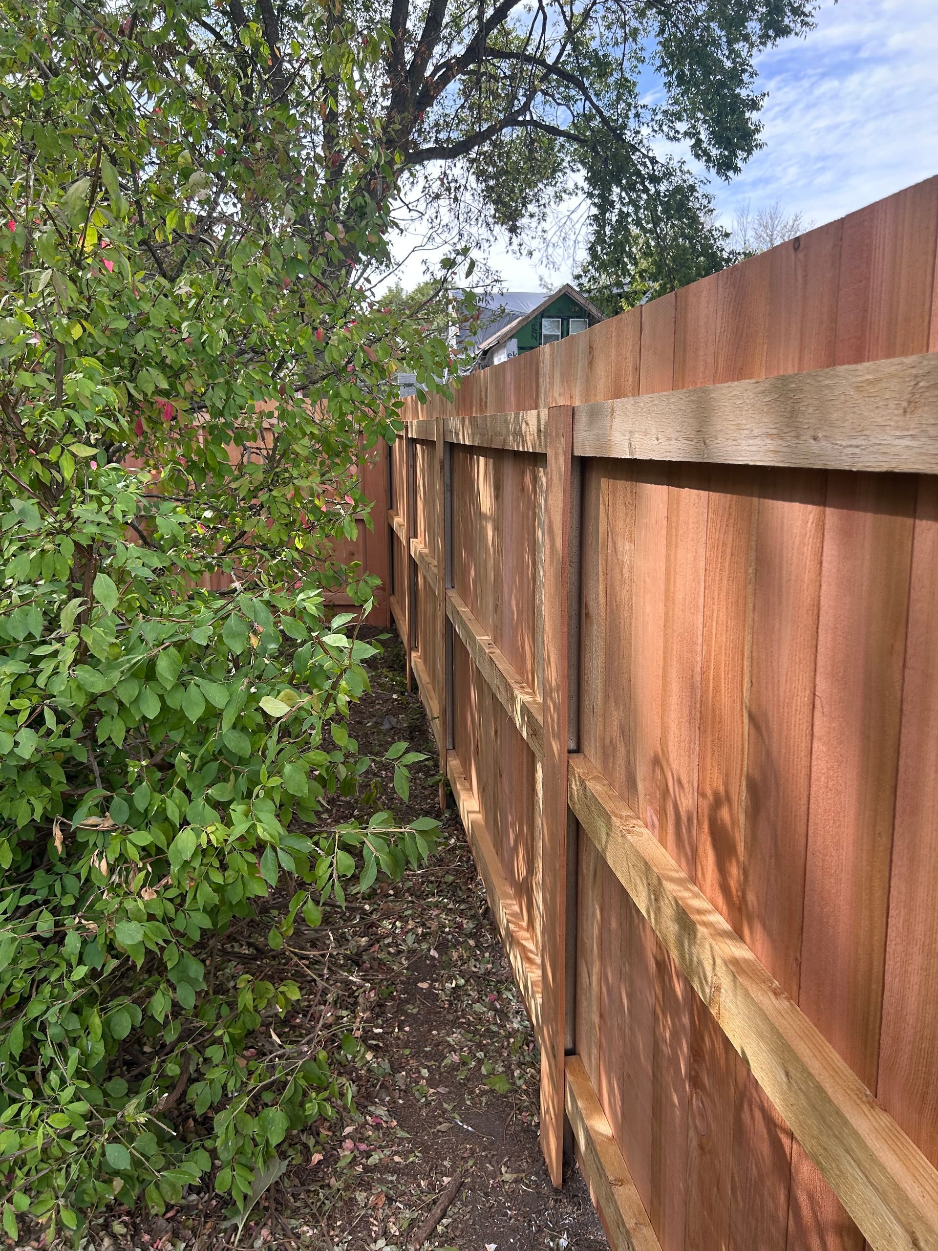 Wooden fence along a narrow dirt path next to green shrubbery under a partly cloudy sky.