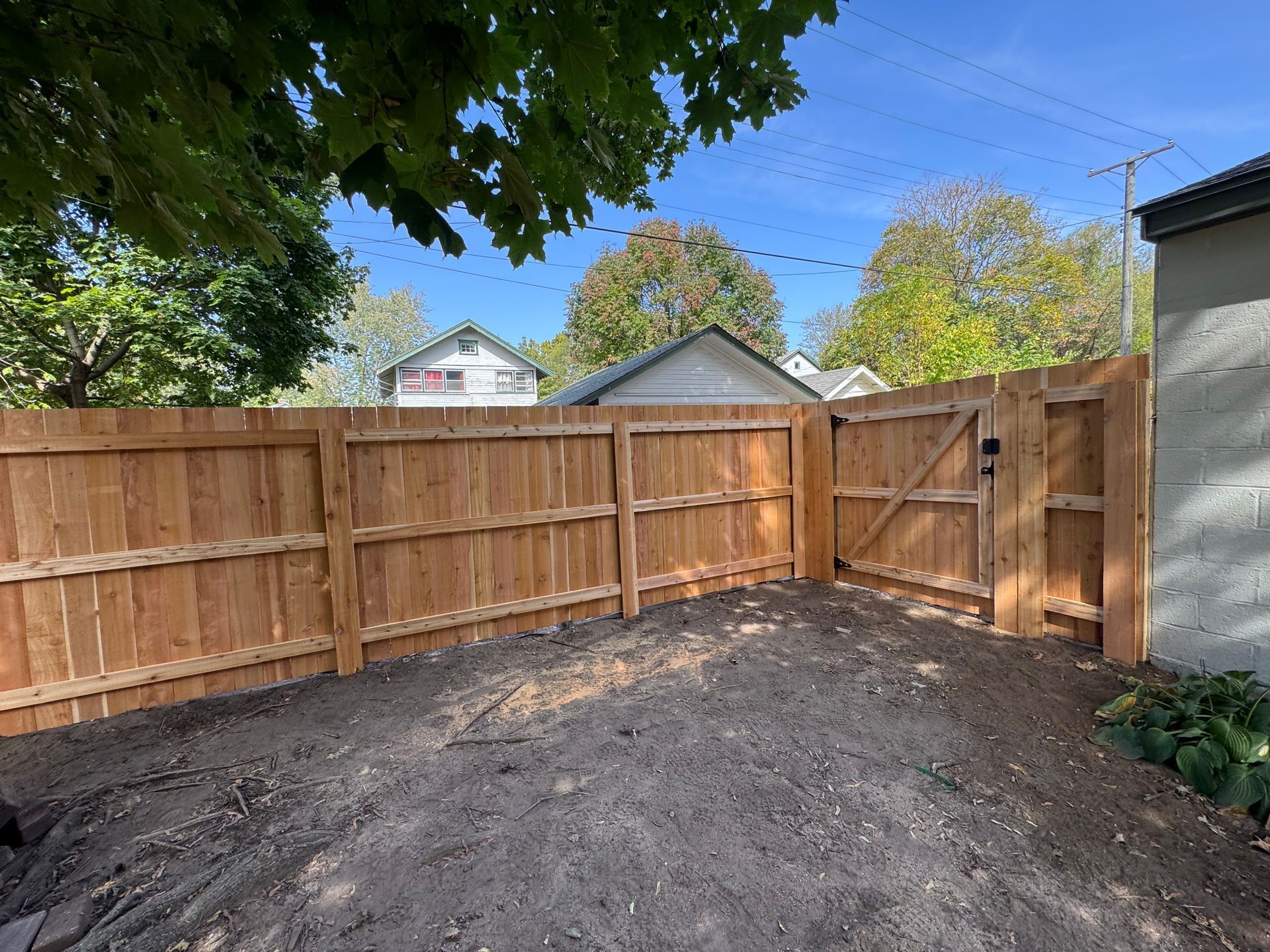 Wooden fence with gate, enclosing a dirt area, houses and trees in background.