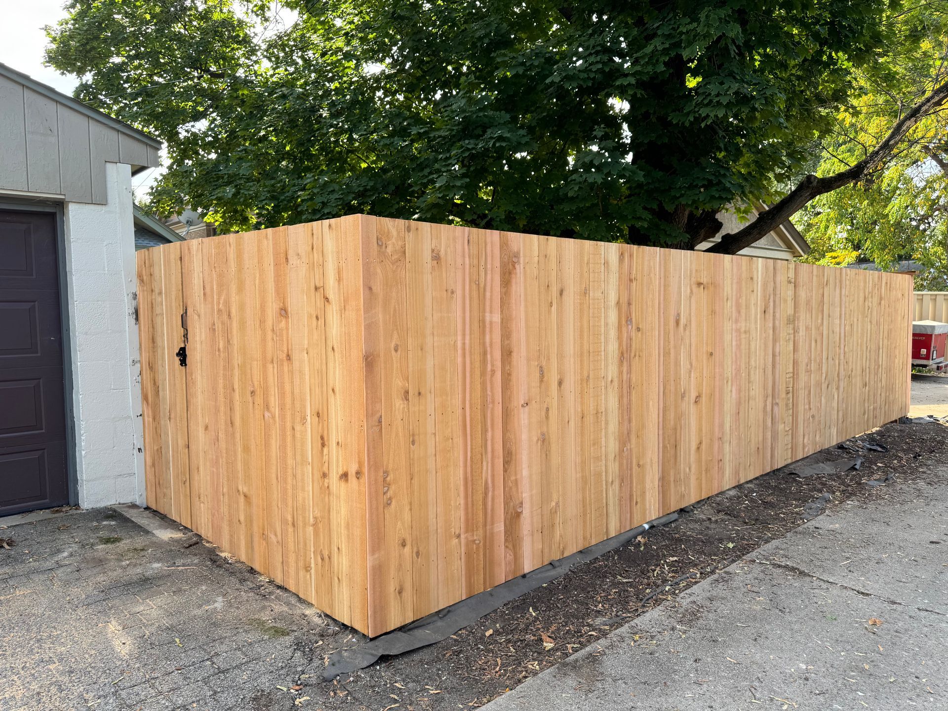Wooden privacy fence surrounding a property, adjacent to a garage and a tree, on a gravel surface.