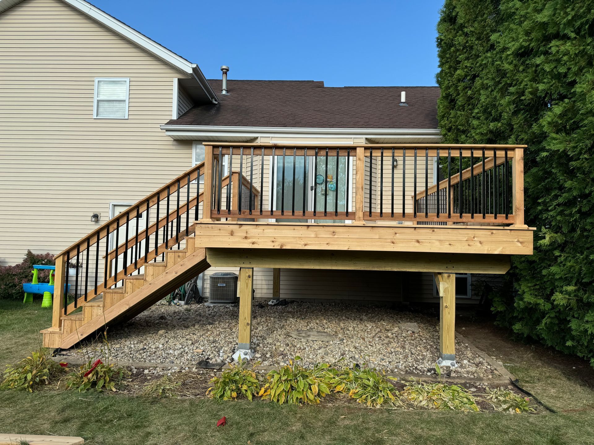 Wooden deck with stairs attached to a beige house, supported by posts on a gravel base.