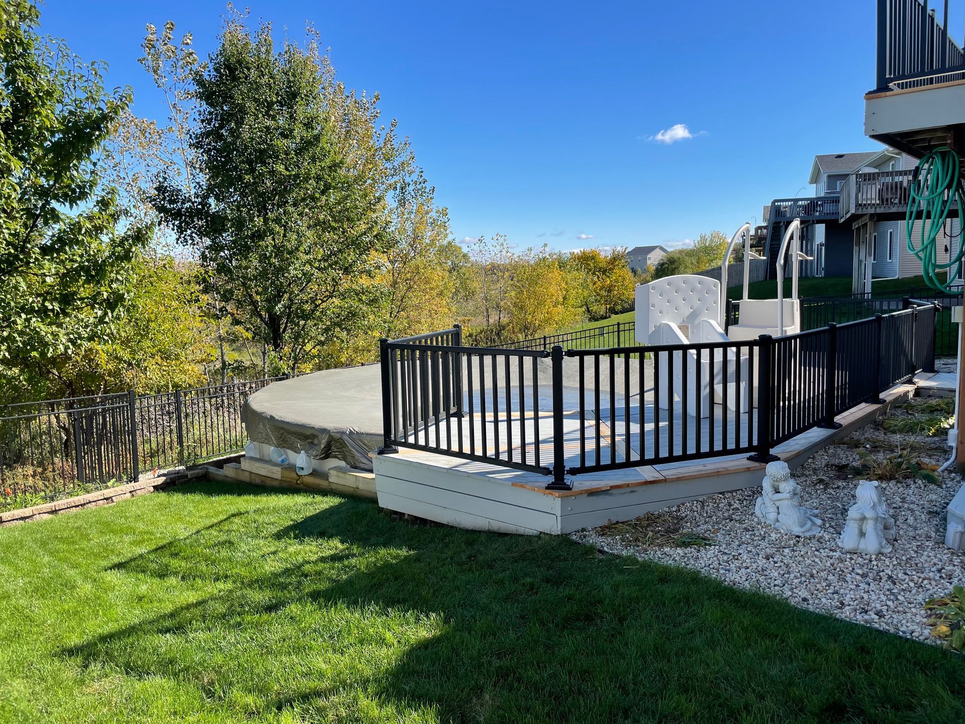 A backyard deck with black railing and concrete surface, next to green grass and trees under a blue sky.