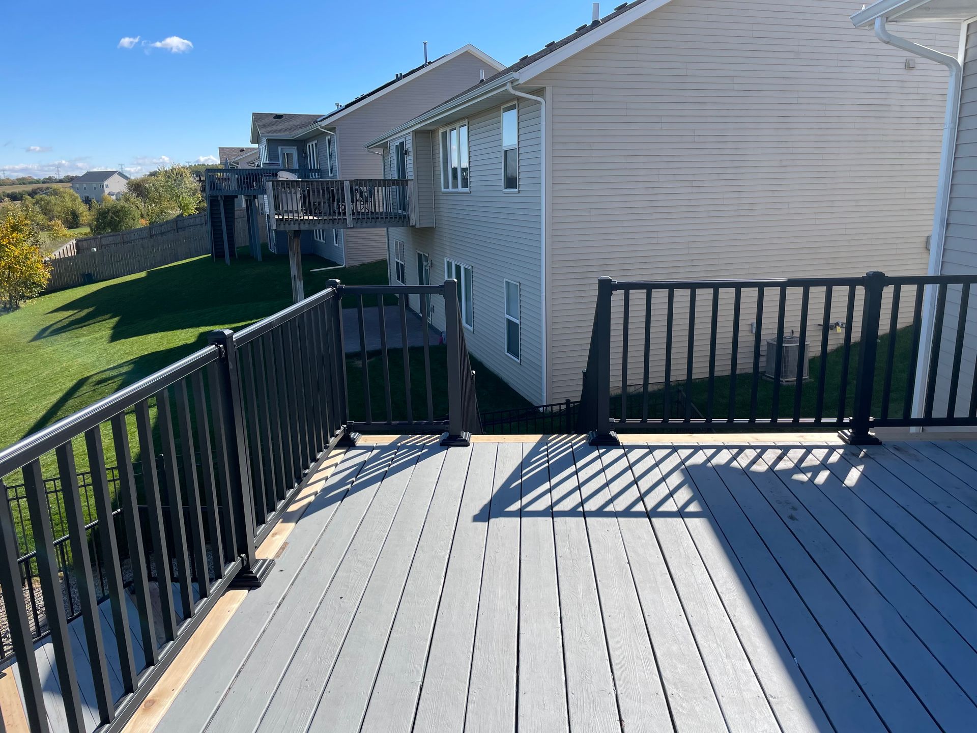 Gray composite deck with black railing next to a house with a grass lawn on a sunny day.