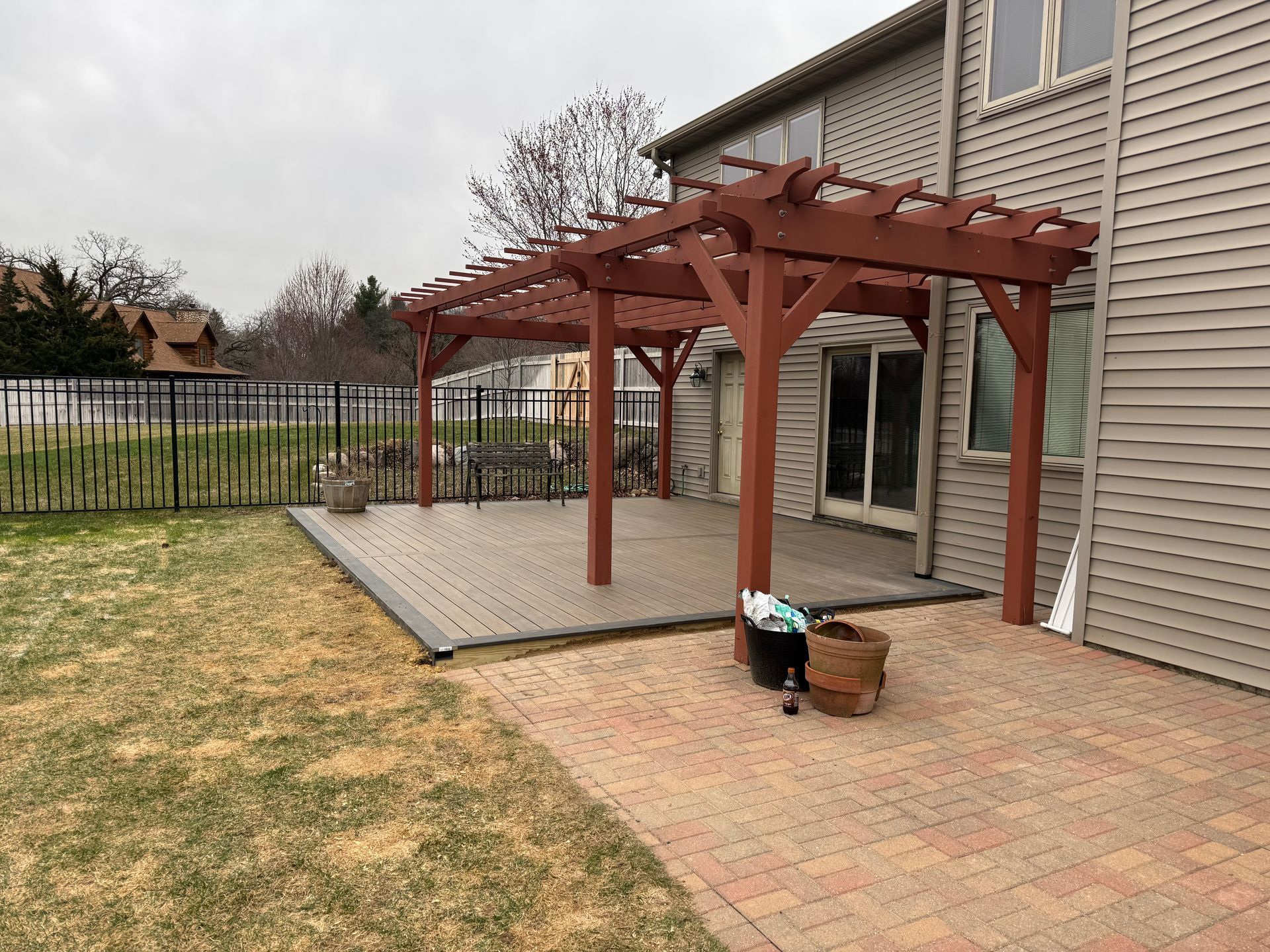 Red pergola over a composite deck next to a brick patio, attached to a two-story beige house.