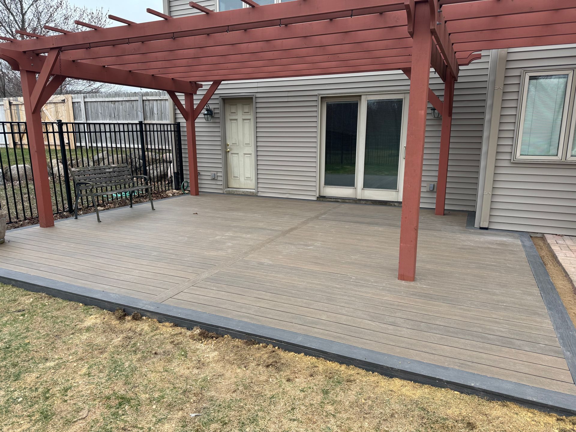 Red pergola over a concrete patio next to a house with a sliding glass door and a black metal fence.